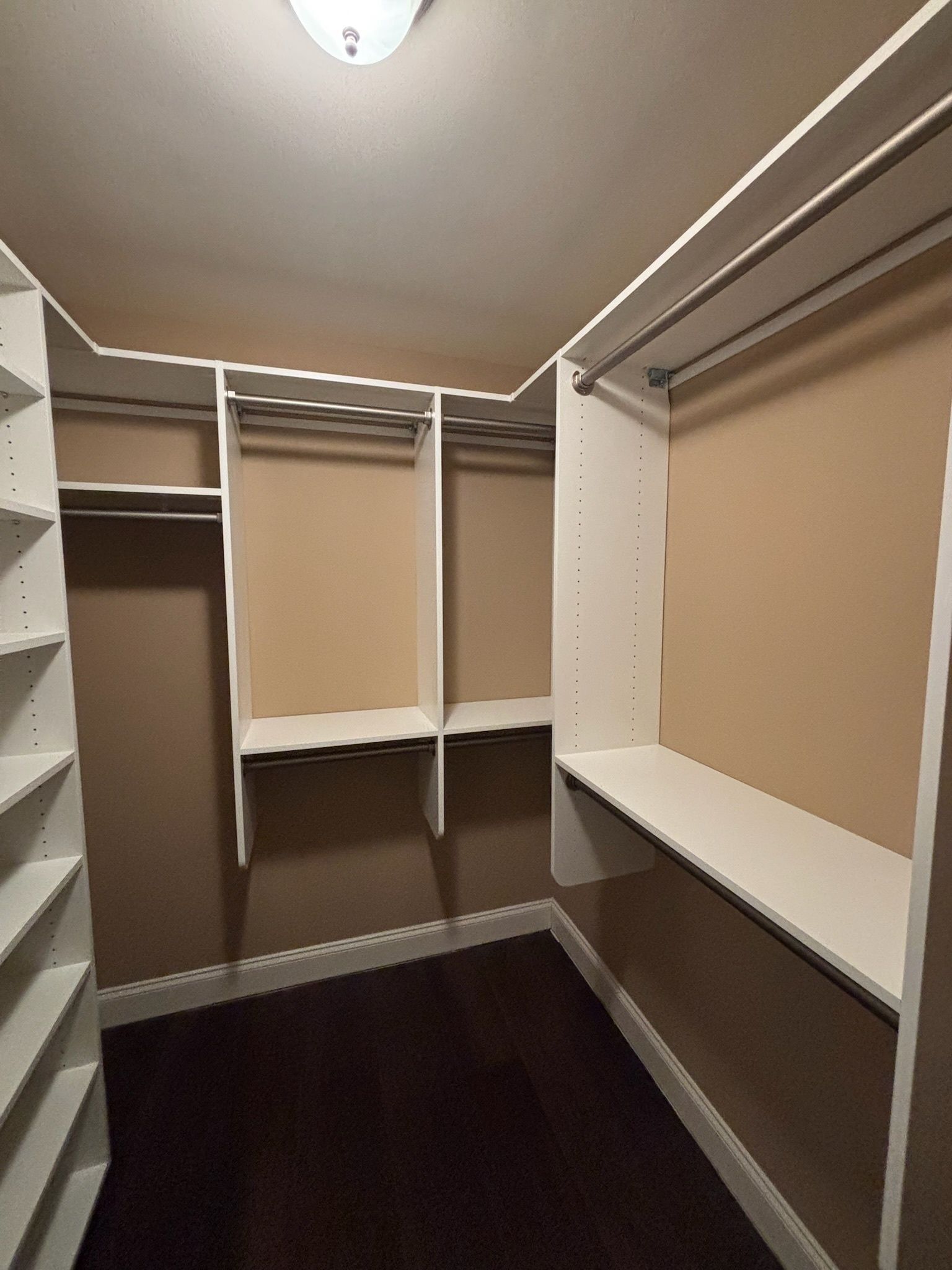 A walk-in closet with white shelving, hanging rods, and dark wood floors against tan walls.