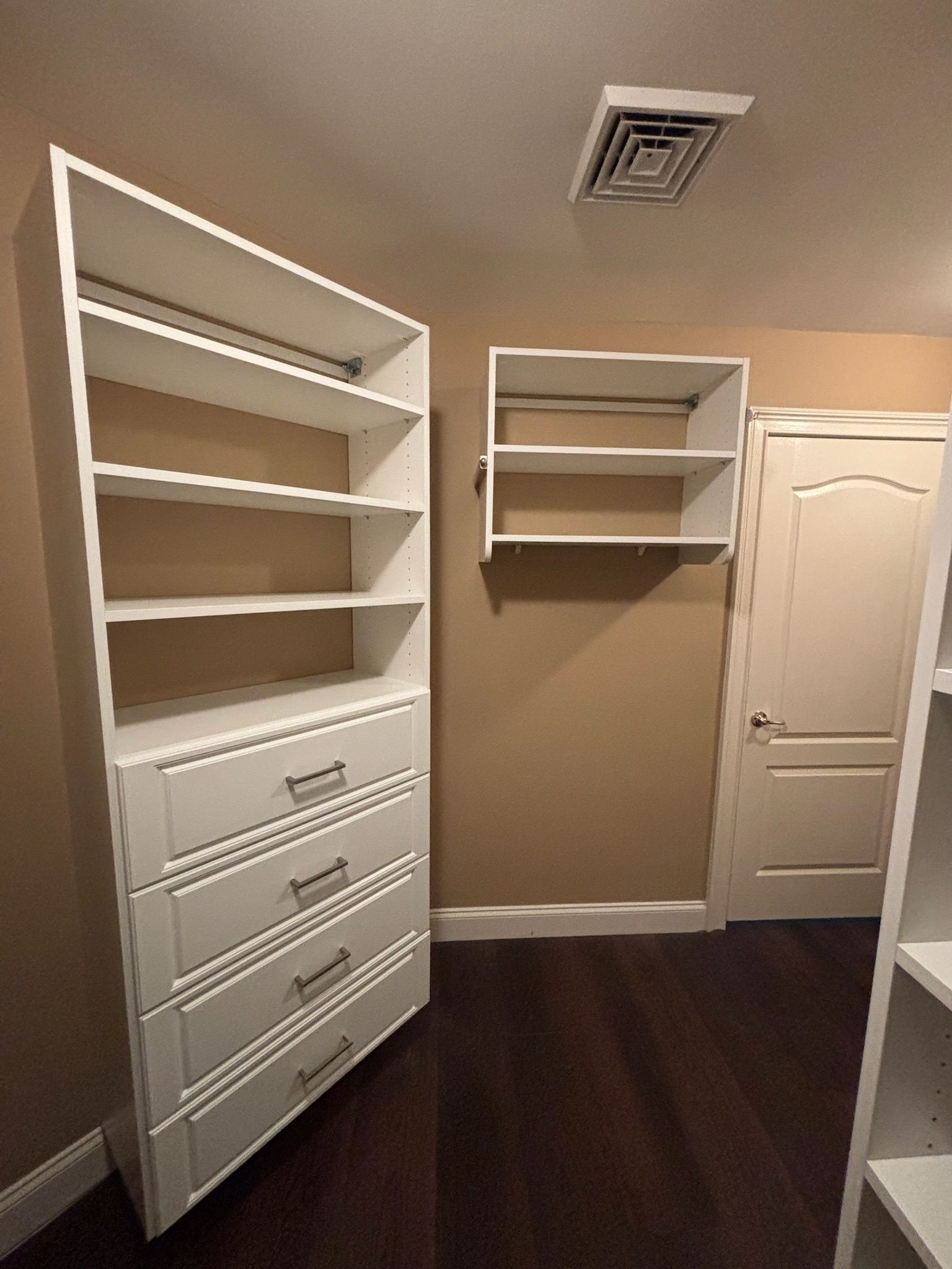White shelving units and a four-drawer dresser installed on a beige wall in a closet with dark wood flooring.
