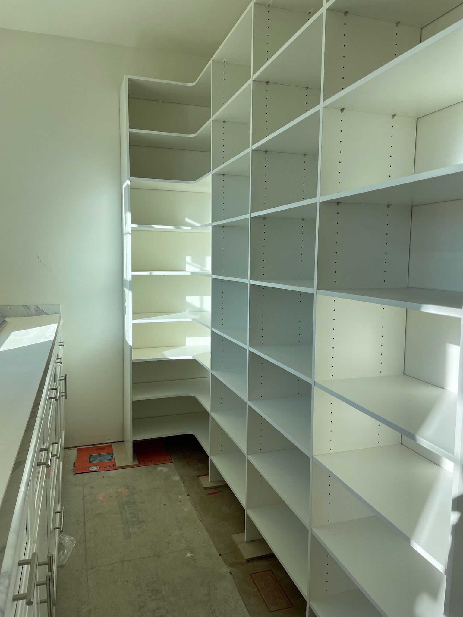 Empty white walk-in closet shelving with corner storage units and a counter on the left in a room with unfinished floors.