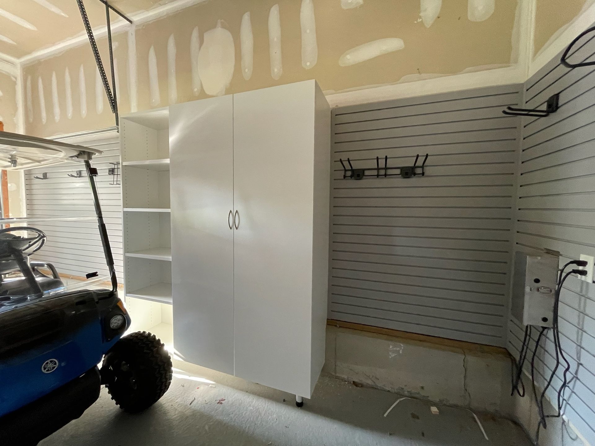 A white storage cabinet and shelving unit in a garage with grey slat walls, a golf cart, and visible wall hooks.