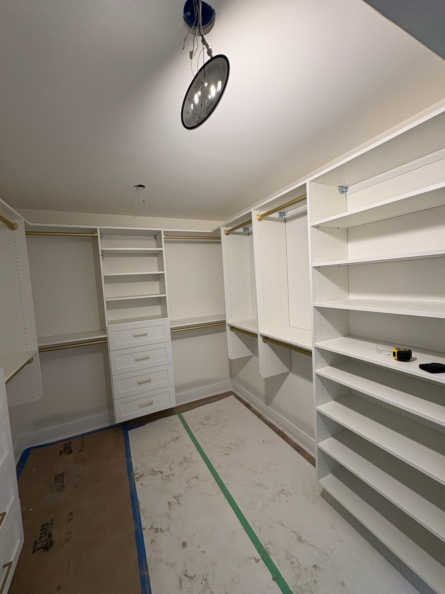 A white walk-in closet under construction with open shelves, hanging rods, and drawers, featuring a central light fixture.