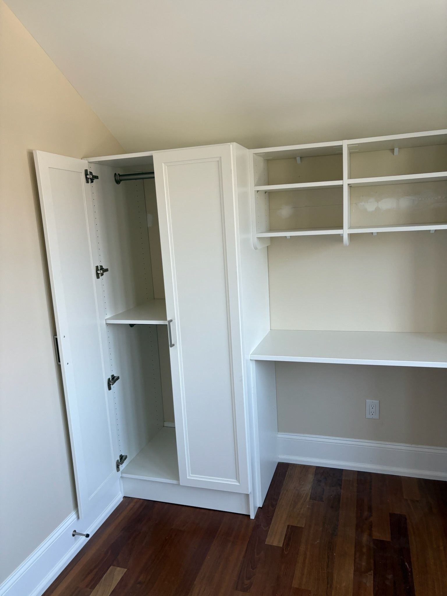A white built-in wardrobe with an open door next to a desk area with two open shelves against a light wall and wood floor.