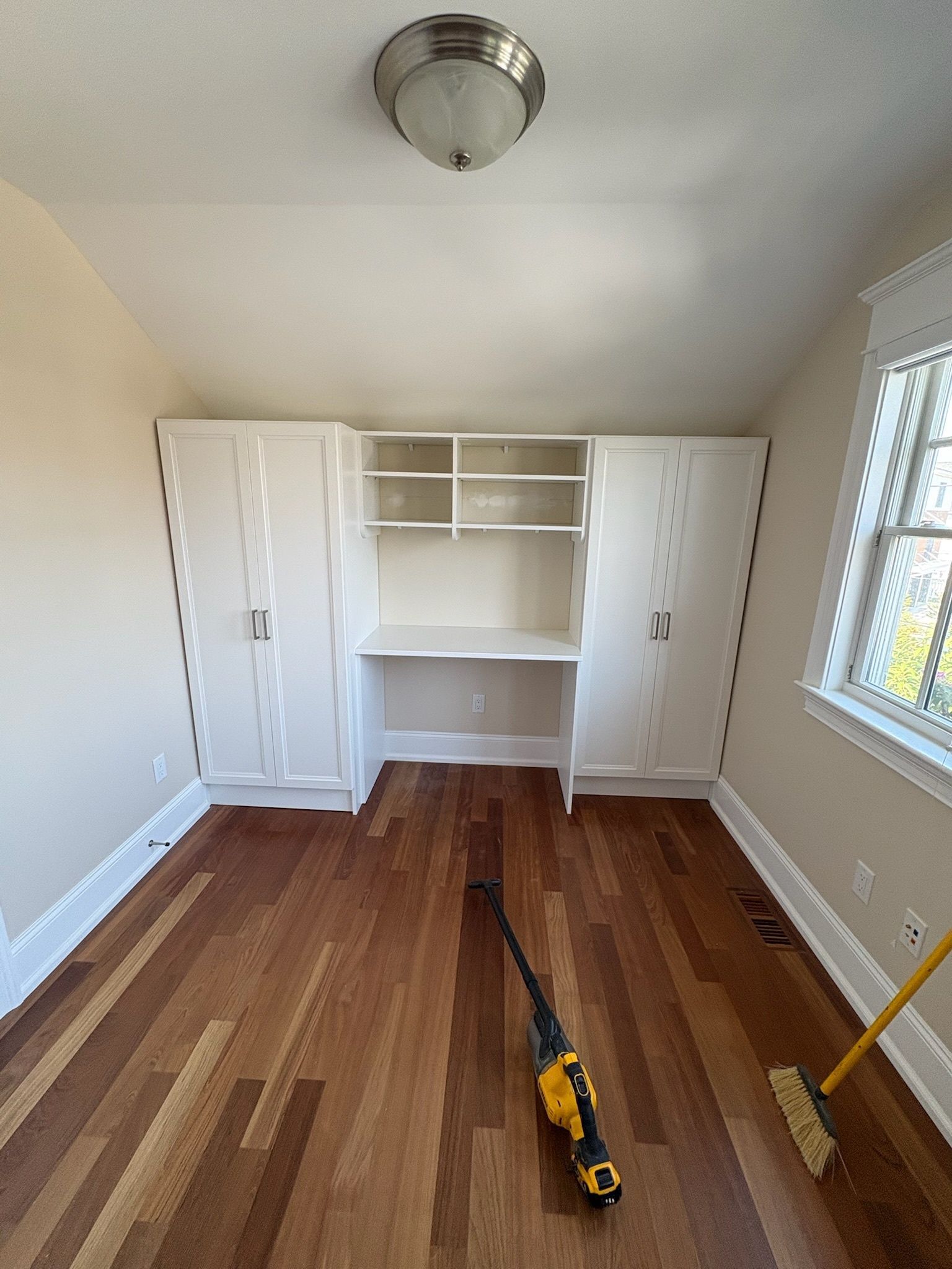 A room with white built-in cabinets and a desk, hardwood floors, a ceiling light, and a vacuum cleaner on the floor.