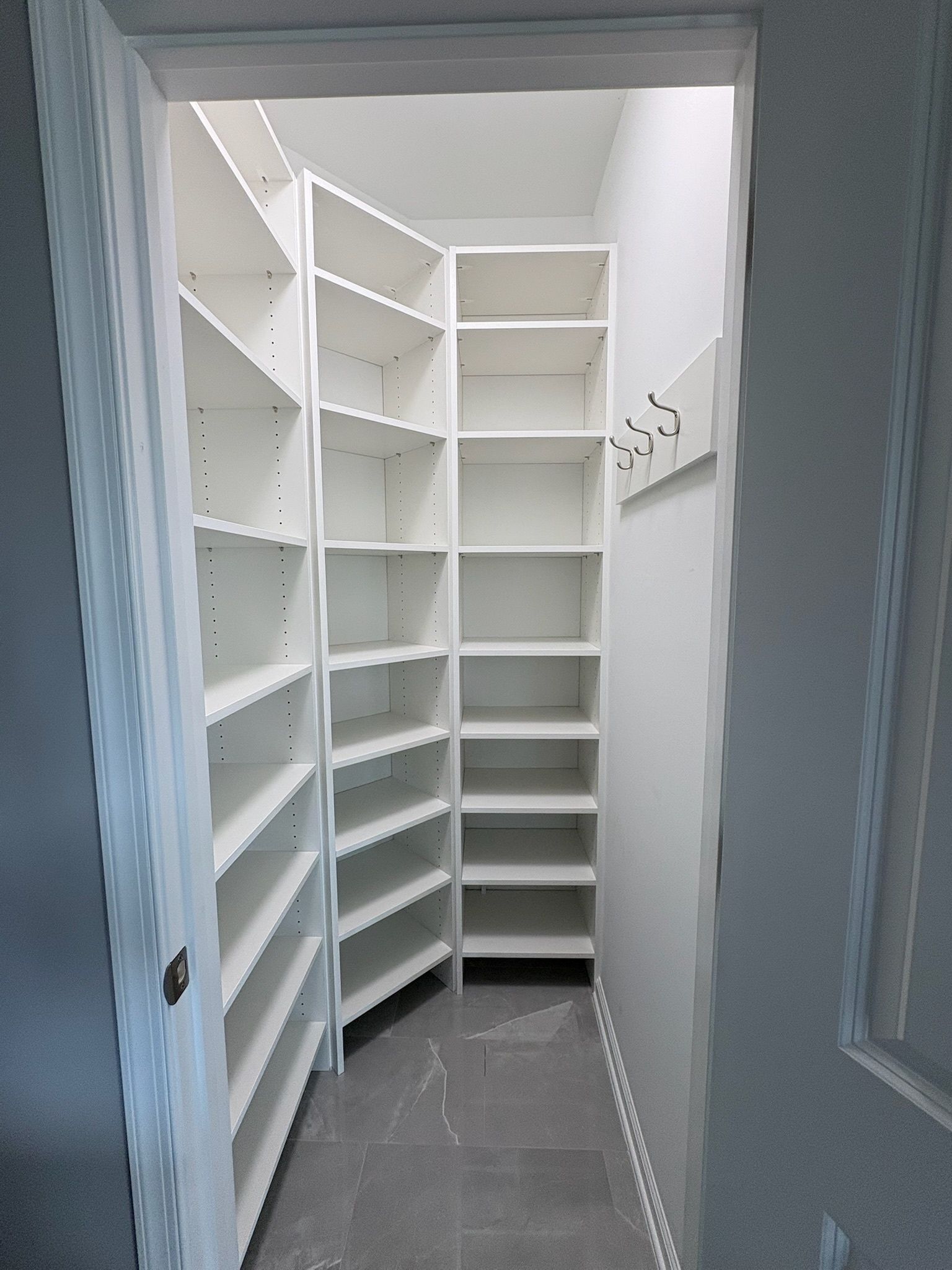 A walk-in pantry with floor-to-ceiling white shelving units along two walls and coat hooks mounted on the right wall.