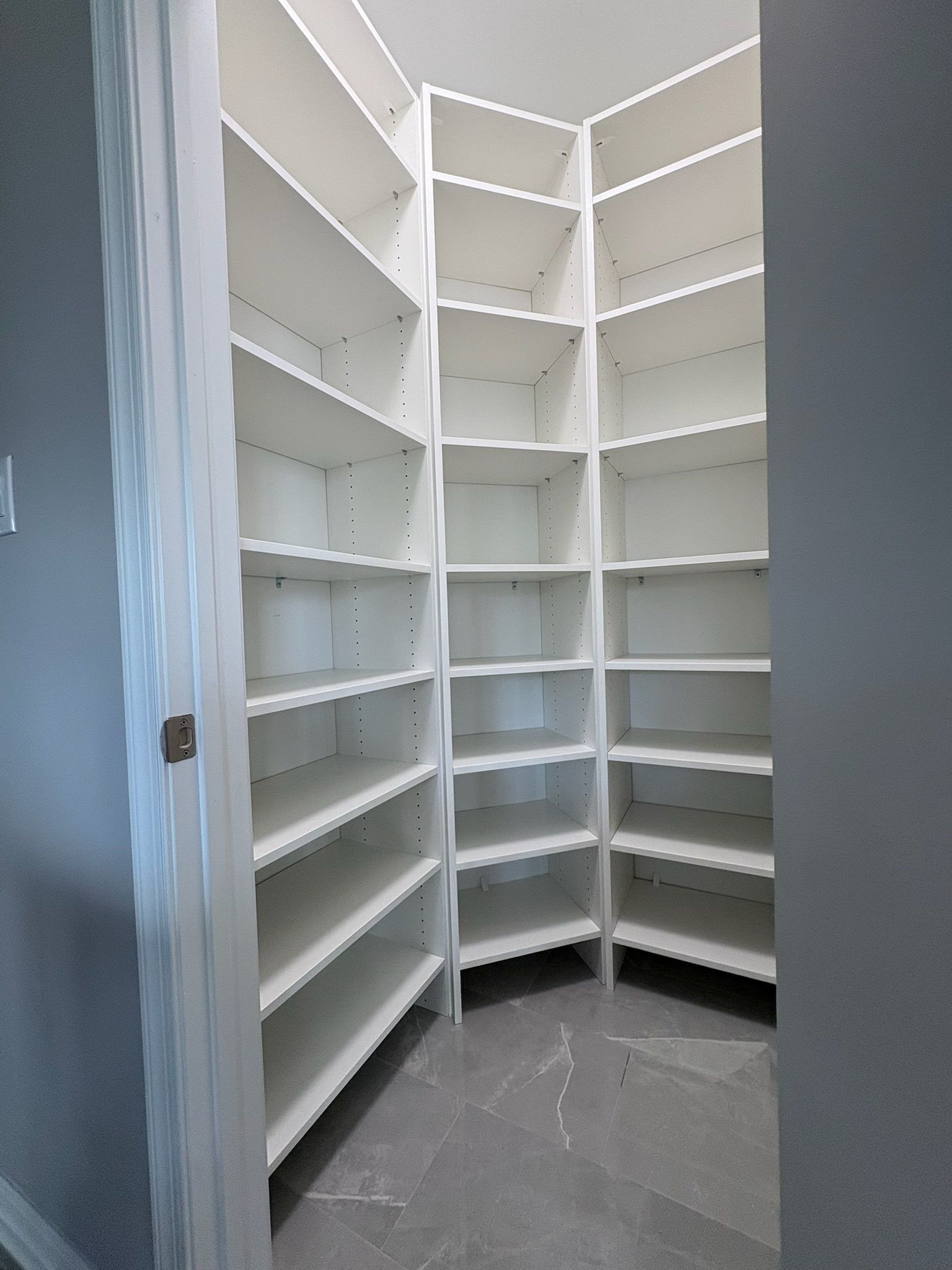 An empty walk-in pantry with white corner shelving units and gray tile flooring.