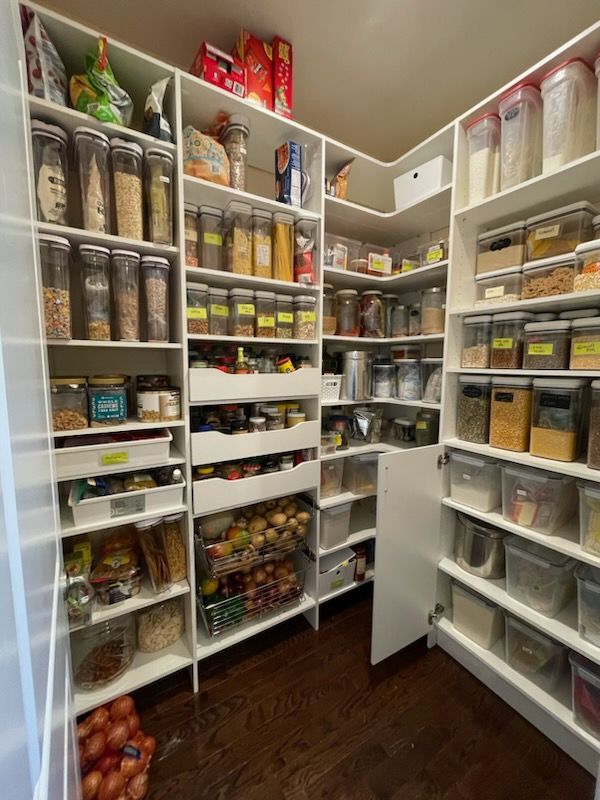 A walk-in pantry with white shelves filled with organized food containers, baskets of produce, and bags on a wood floor.