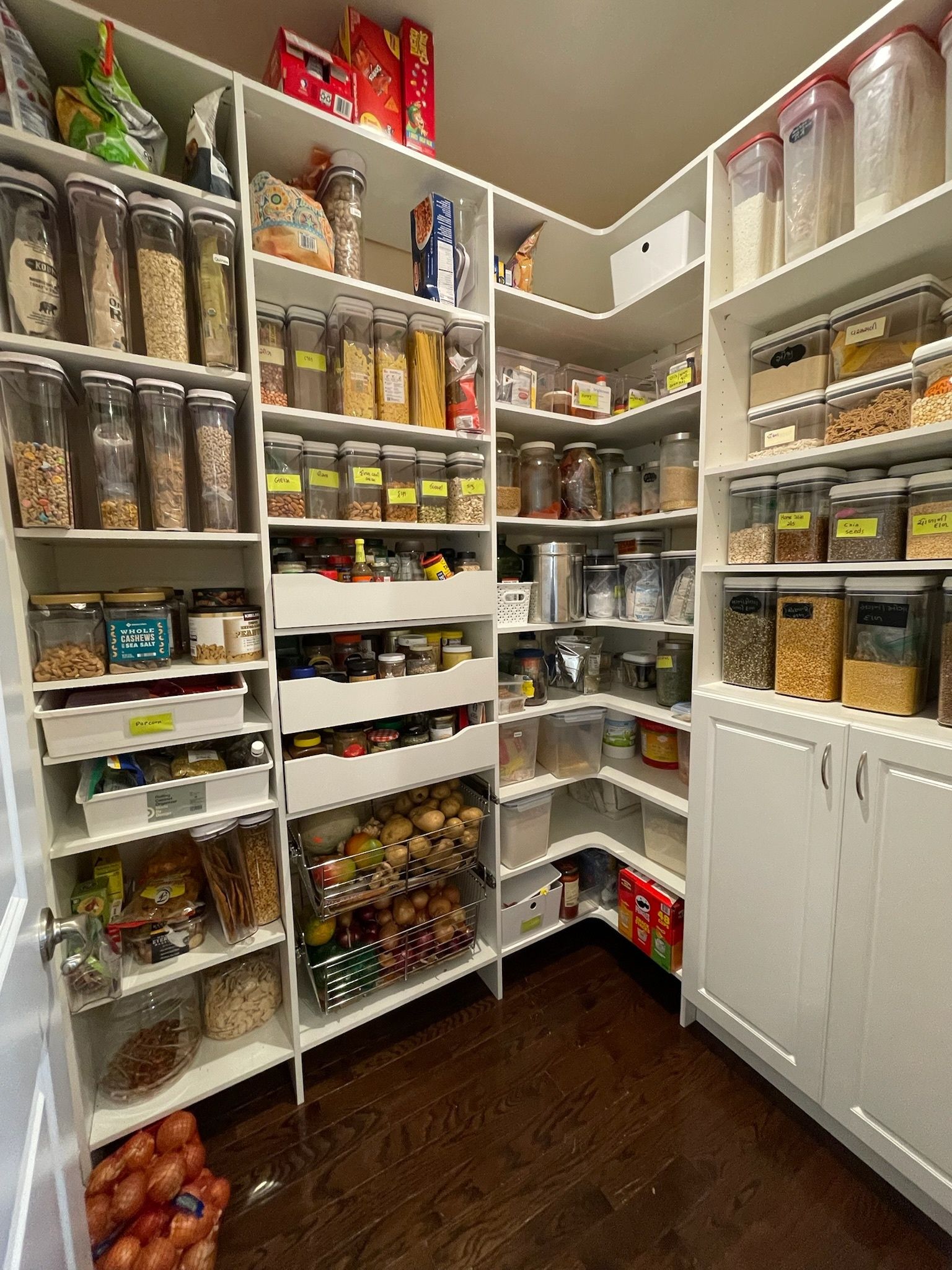 Organized home pantry with white floor-to-ceiling shelves, clear storage containers, and baskets filled with dry goods.