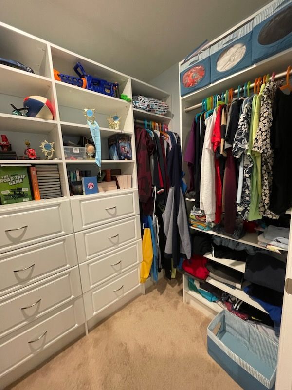 A walk-in closet featuring white wooden shelves, drawers, hanging clothes on racks, and storage bins on the top shelf.