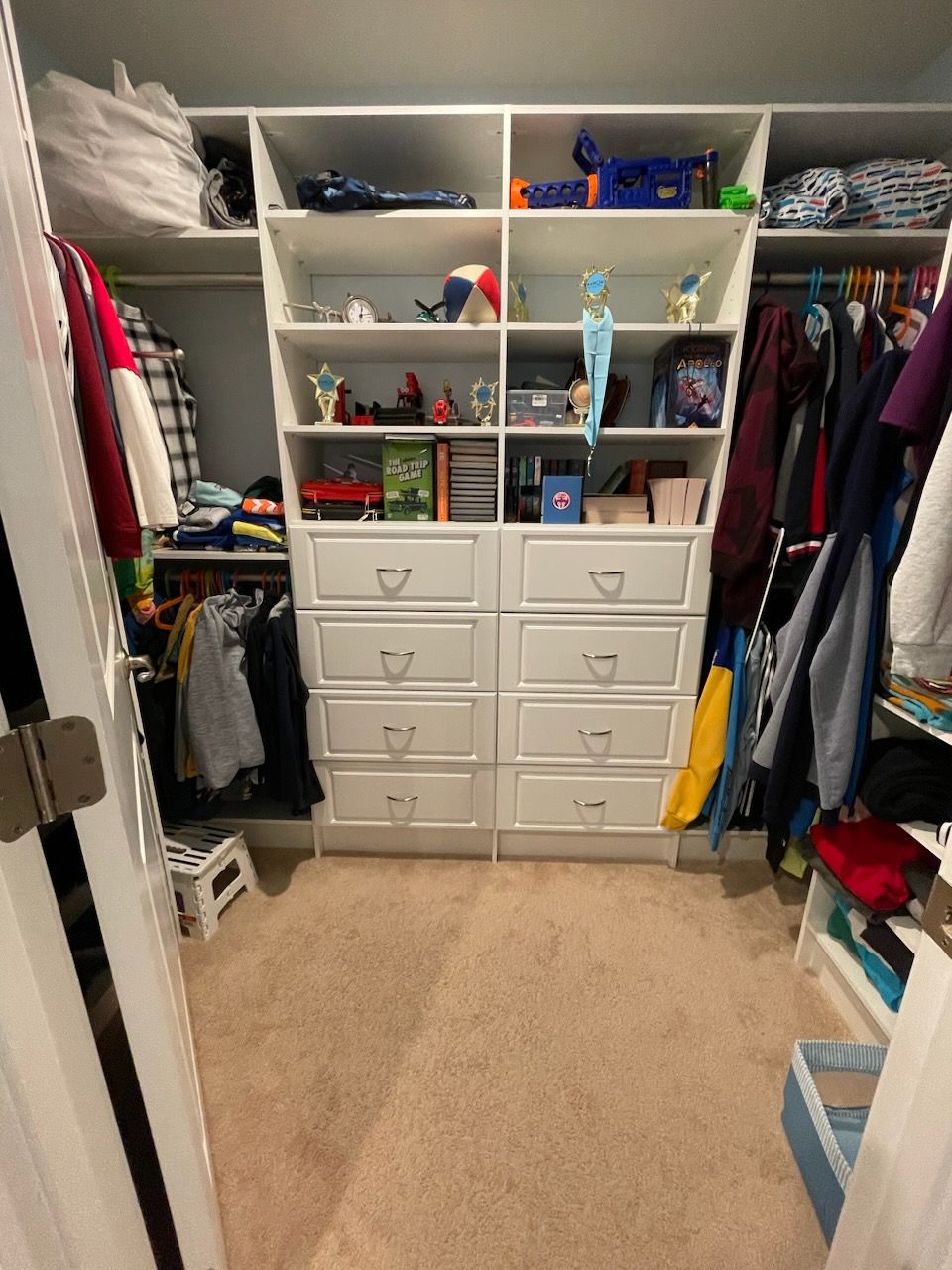 A white walk-in closet with shelves, hanging clothes on both sides, and eight central drawers, carpeted throughout.