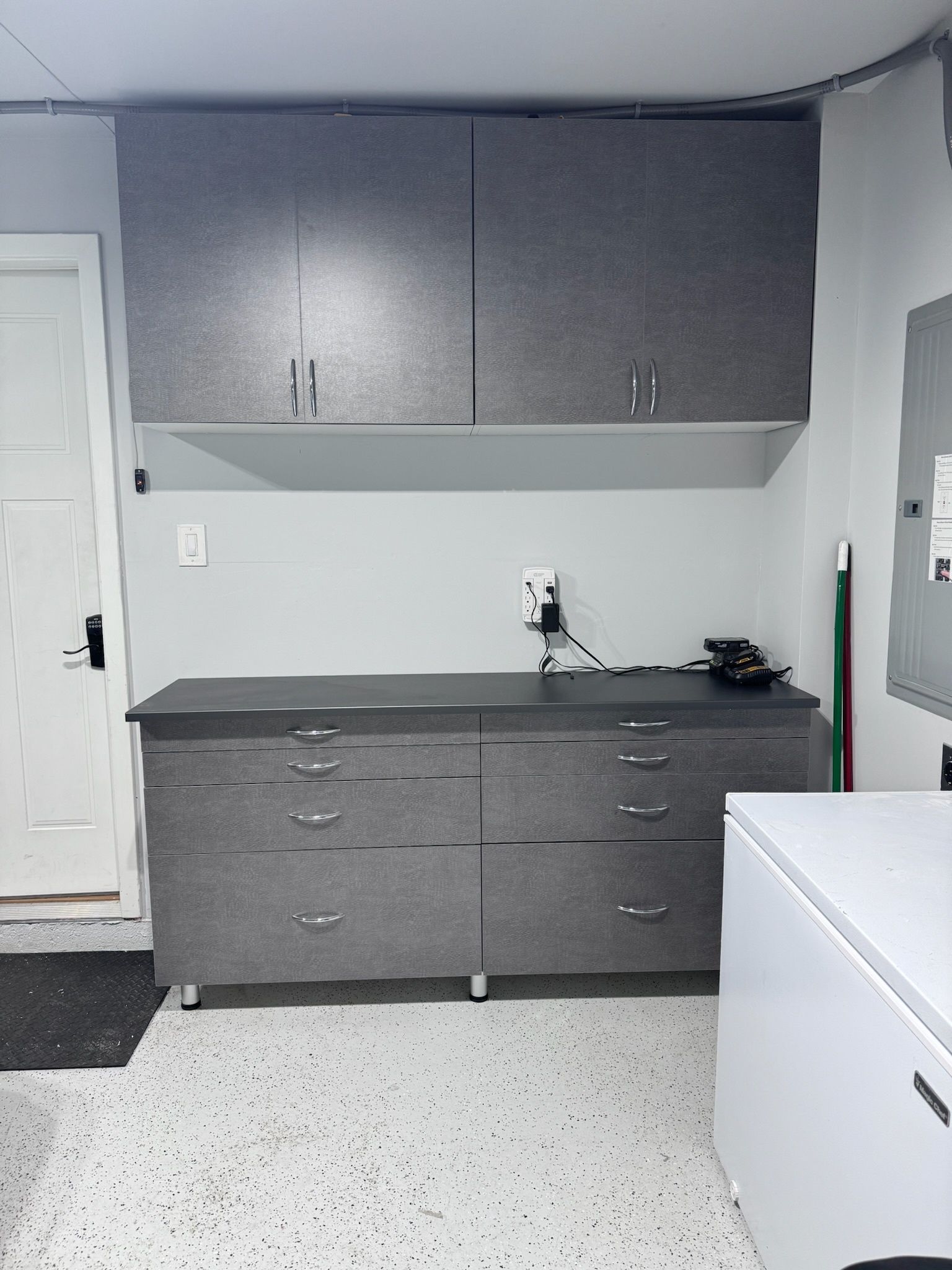 Gray cabinetry with a workbench and overhead storage in a garage with a white speckled floor and a nearby chest freezer.