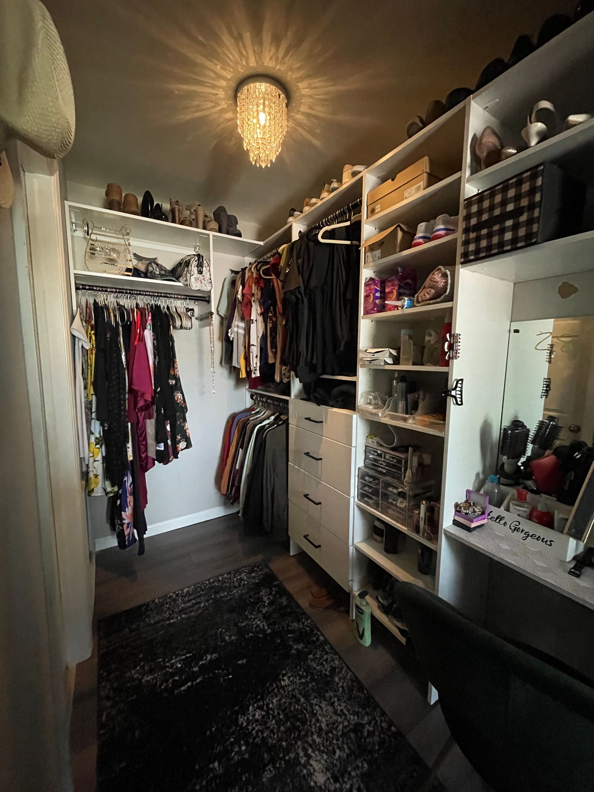 A walk-in closet featuring hanging racks, open shelving for shoes and accessories, a vanity area, and a dark rug.