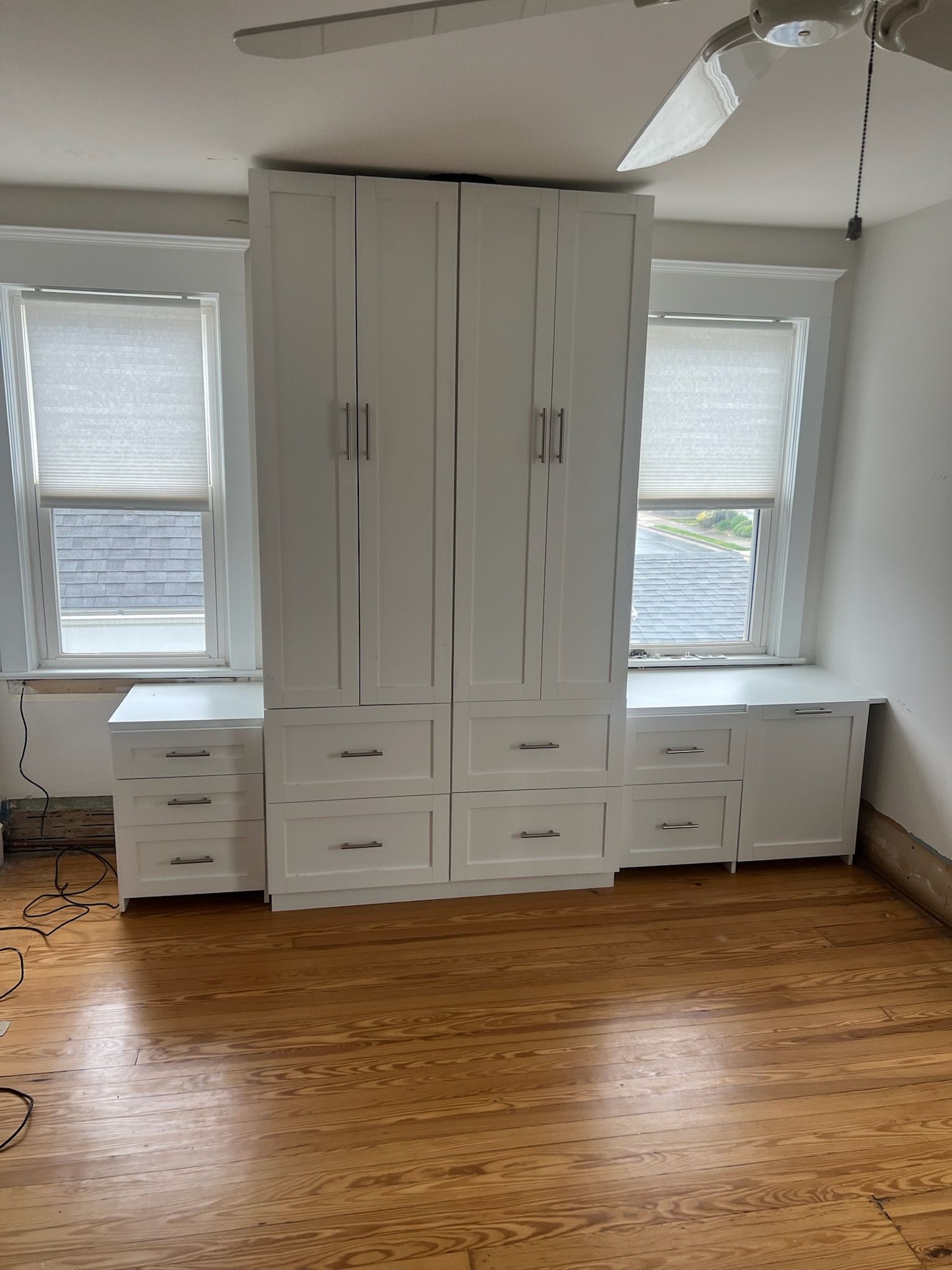 White storage cabinetry featuring central tall wardrobes and flanking lower drawers, situated between two windows.