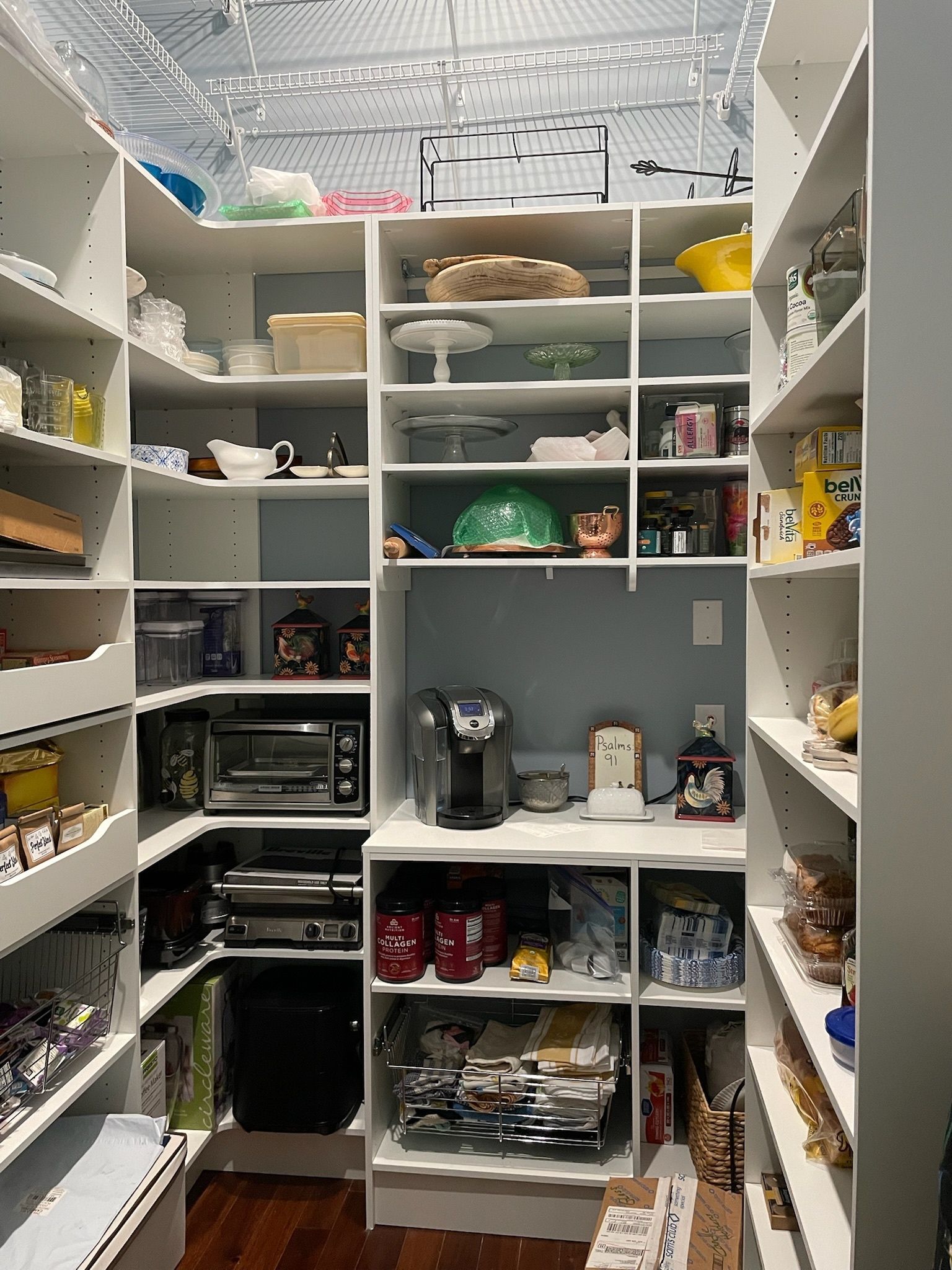 A white walk-in pantry with wrap-around shelves filled with kitchen appliances, dishware, and various food items.