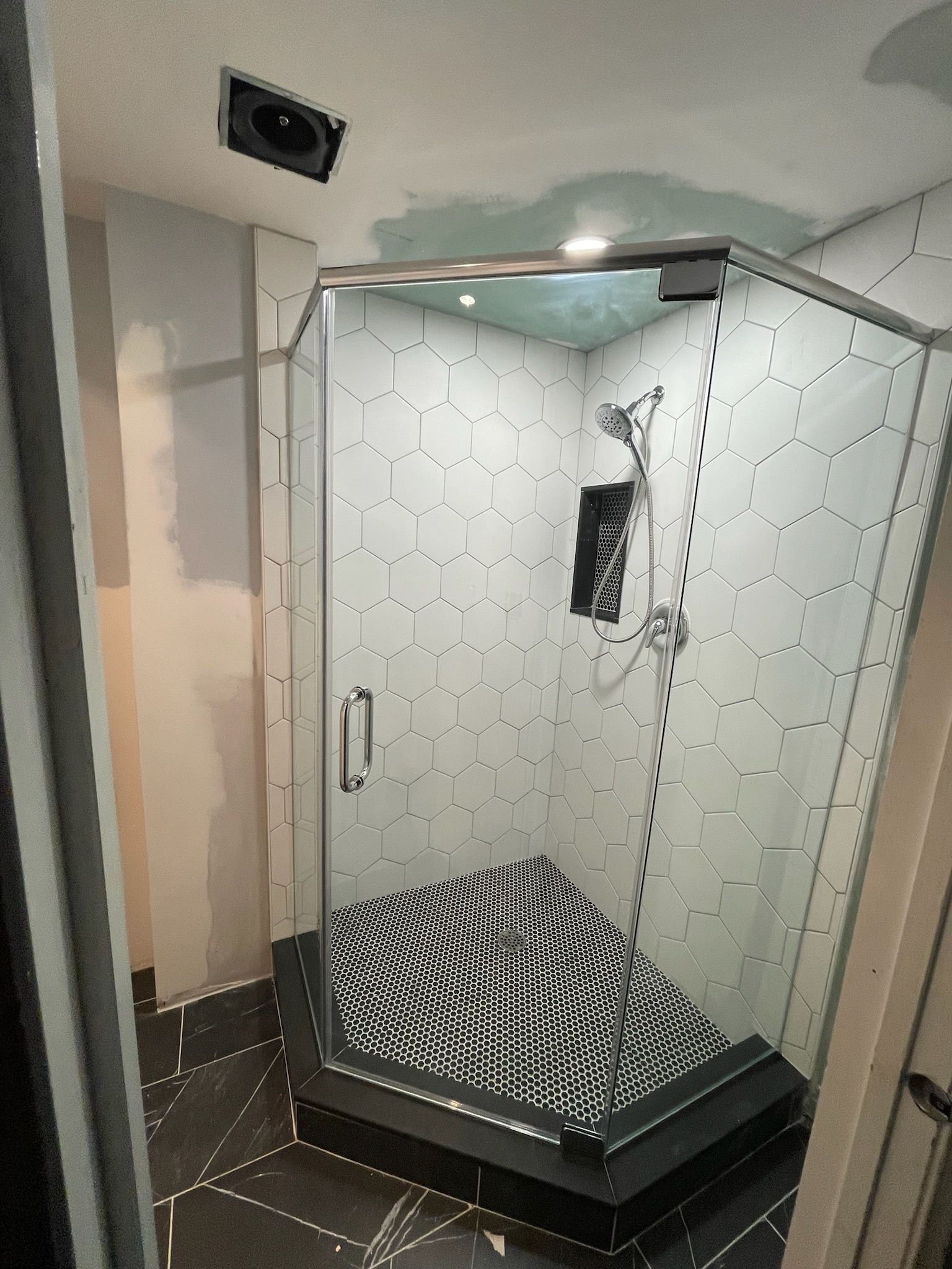 A modern corner shower with white hexagonal wall tiles, a black geometric floor pattern, and glass doors.
