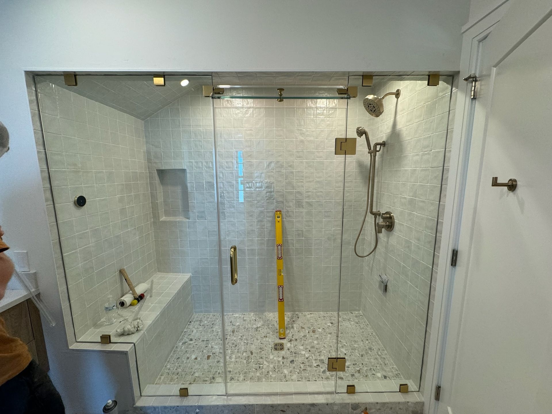 A walk-in shower with white subway tile walls, a built-in bench, a glass door, and brass hardware in a bathroom.