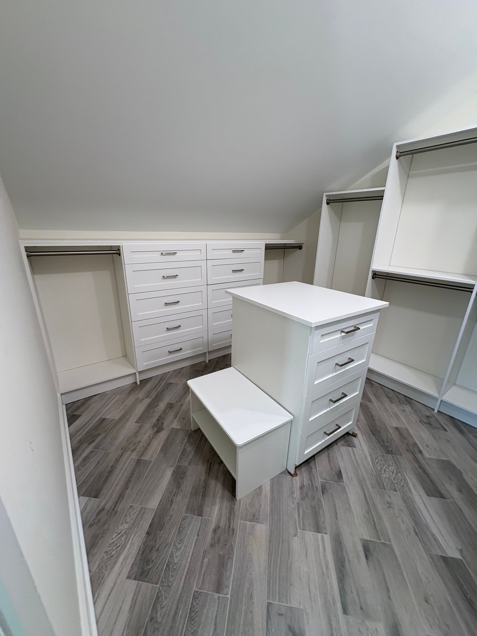 A white walk-in closet with built-in drawers, hanging rods, and a matching center island with a small step stool.