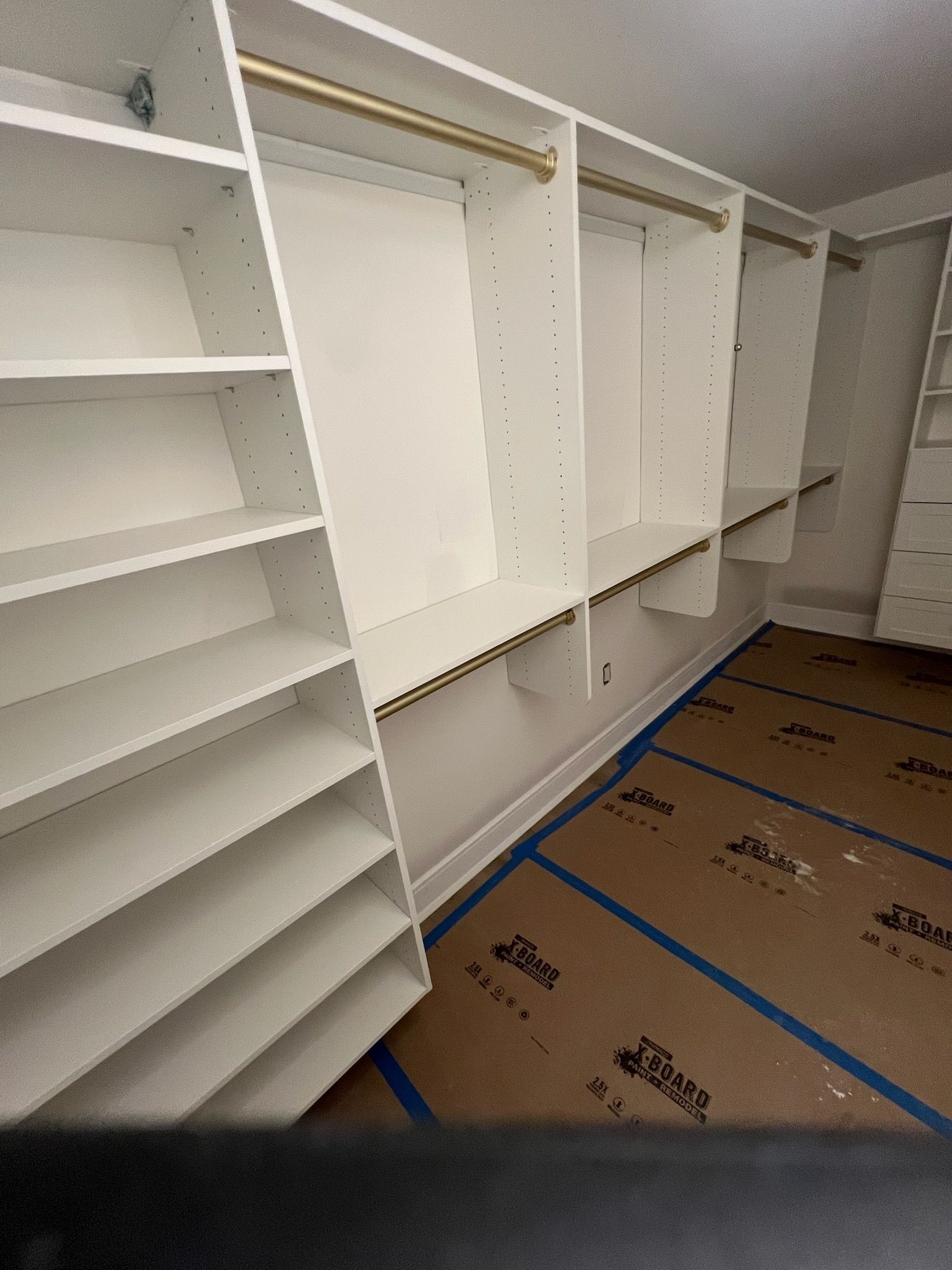 A white, custom-built walk-in closet system with open shelving and gold hanging rods in a room with floor protection paper.