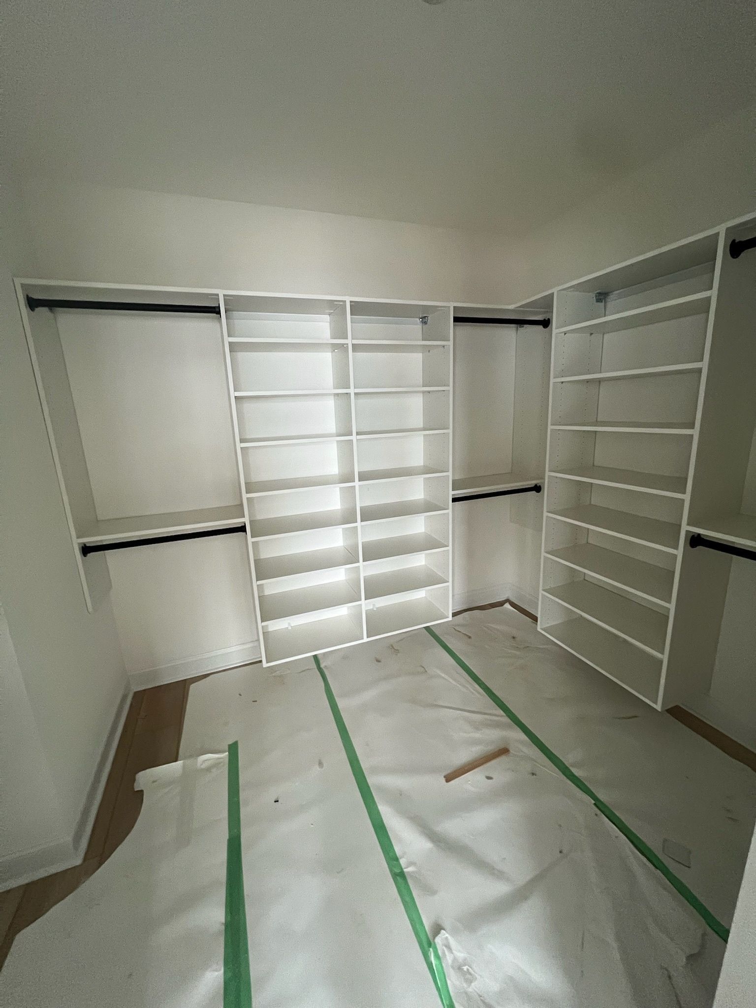 An empty walk-in closet with white shelving units and clothing rods installed along the walls over protective floor paper.