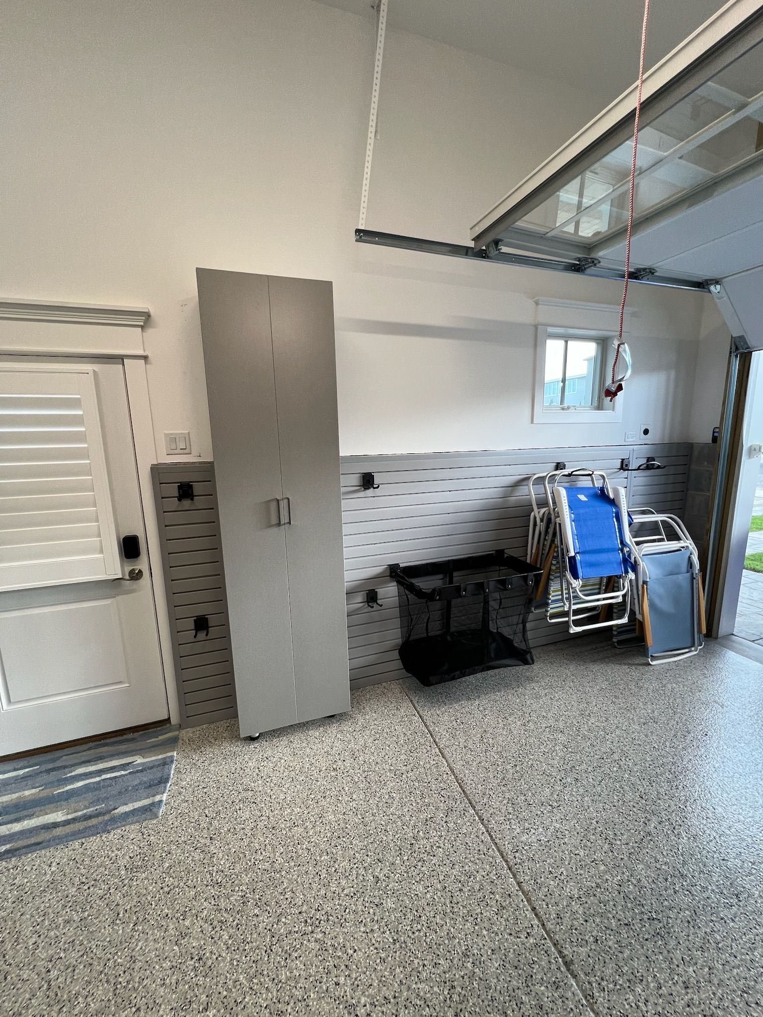 A garage with a gray cabinet, pegboard walls, a black rolling cart, and folded chairs on a speckled concrete floor.