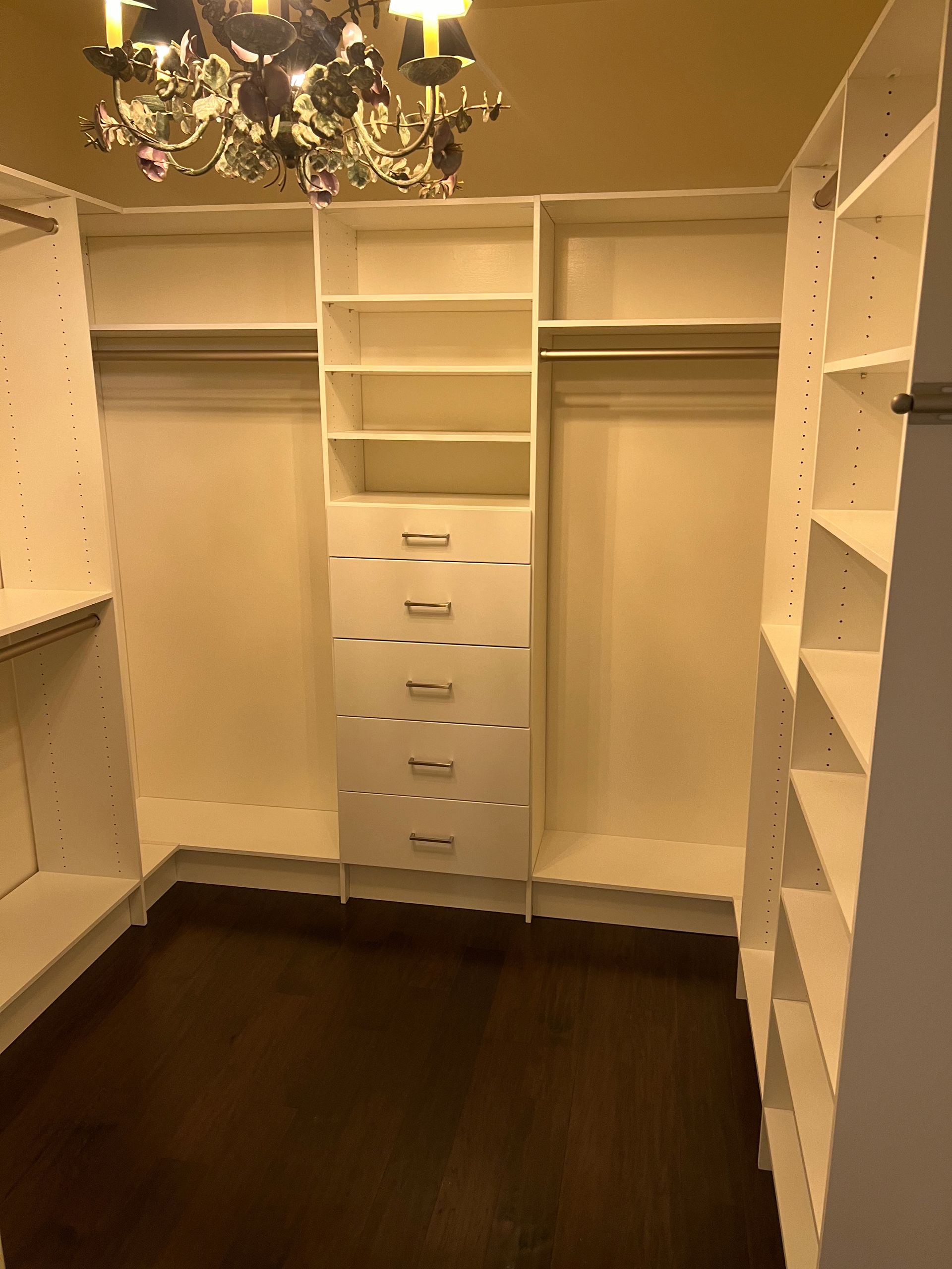 Walk-in closet with white shelving, a central tower of five drawers, hanging rods, and an overhead ornate chandelier.