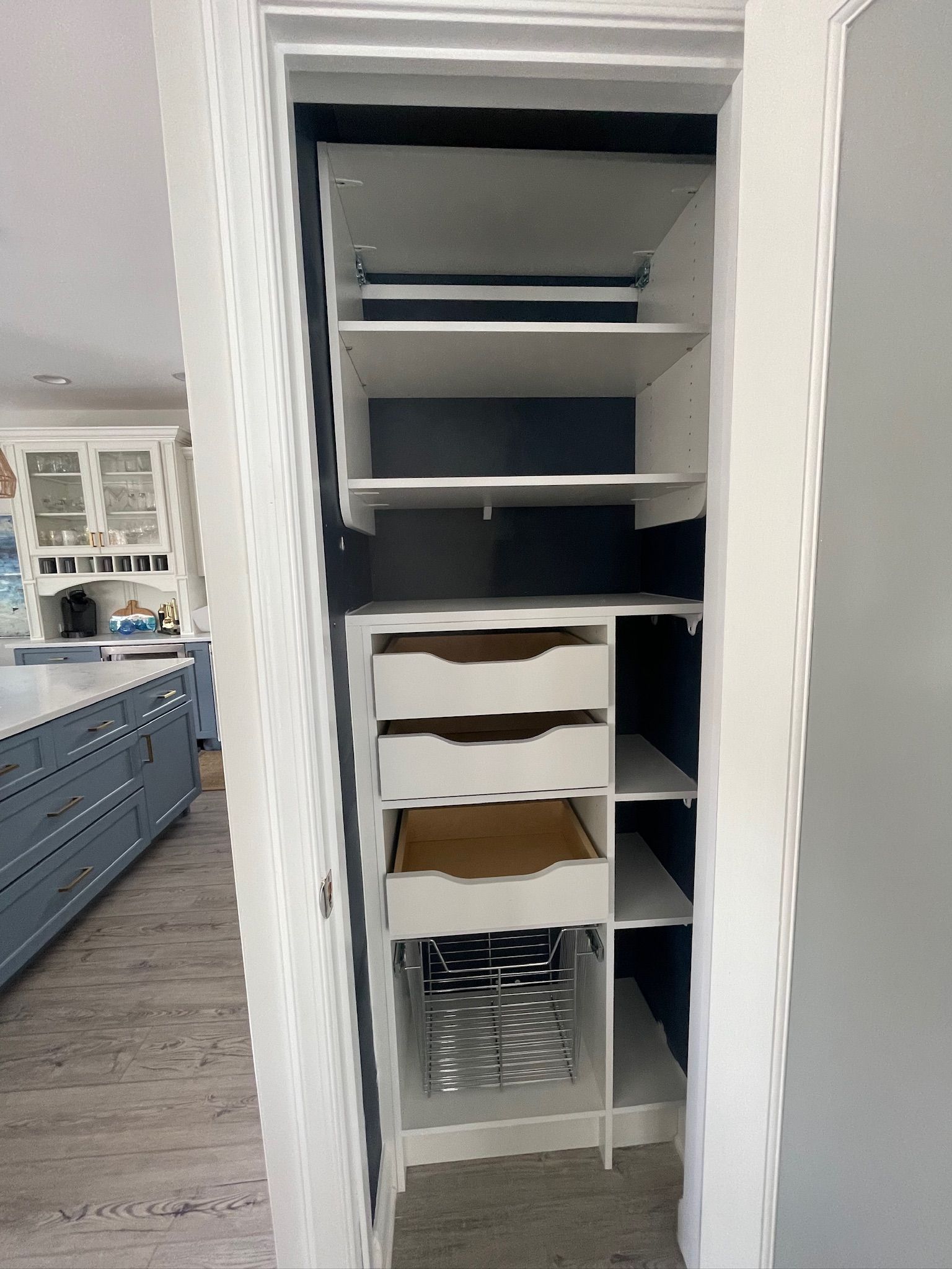 A pantry closet with white shelving and drawers against a dark blue back wall, viewed from a light-colored kitchen.