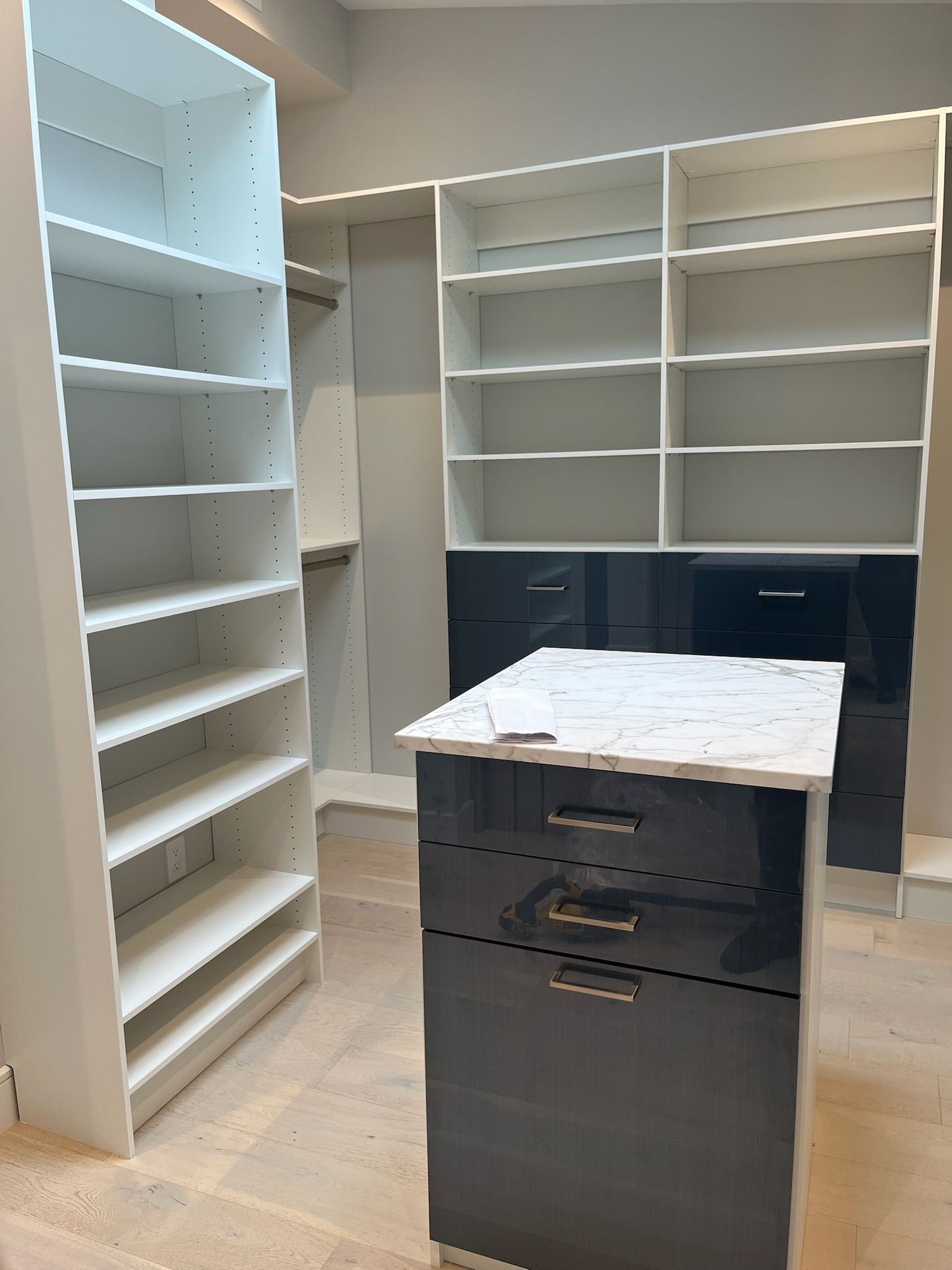 A walk-in closet featuring white shelves, a black dresser island with a marble top, and black drawers under wall shelves.