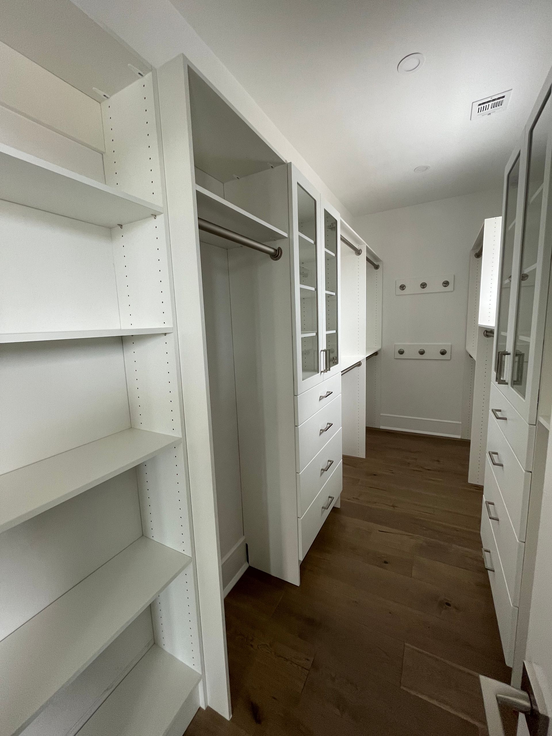 A walk-in closet featuring white modular shelving, a hanging rod, and drawers, set against hardwood flooring.