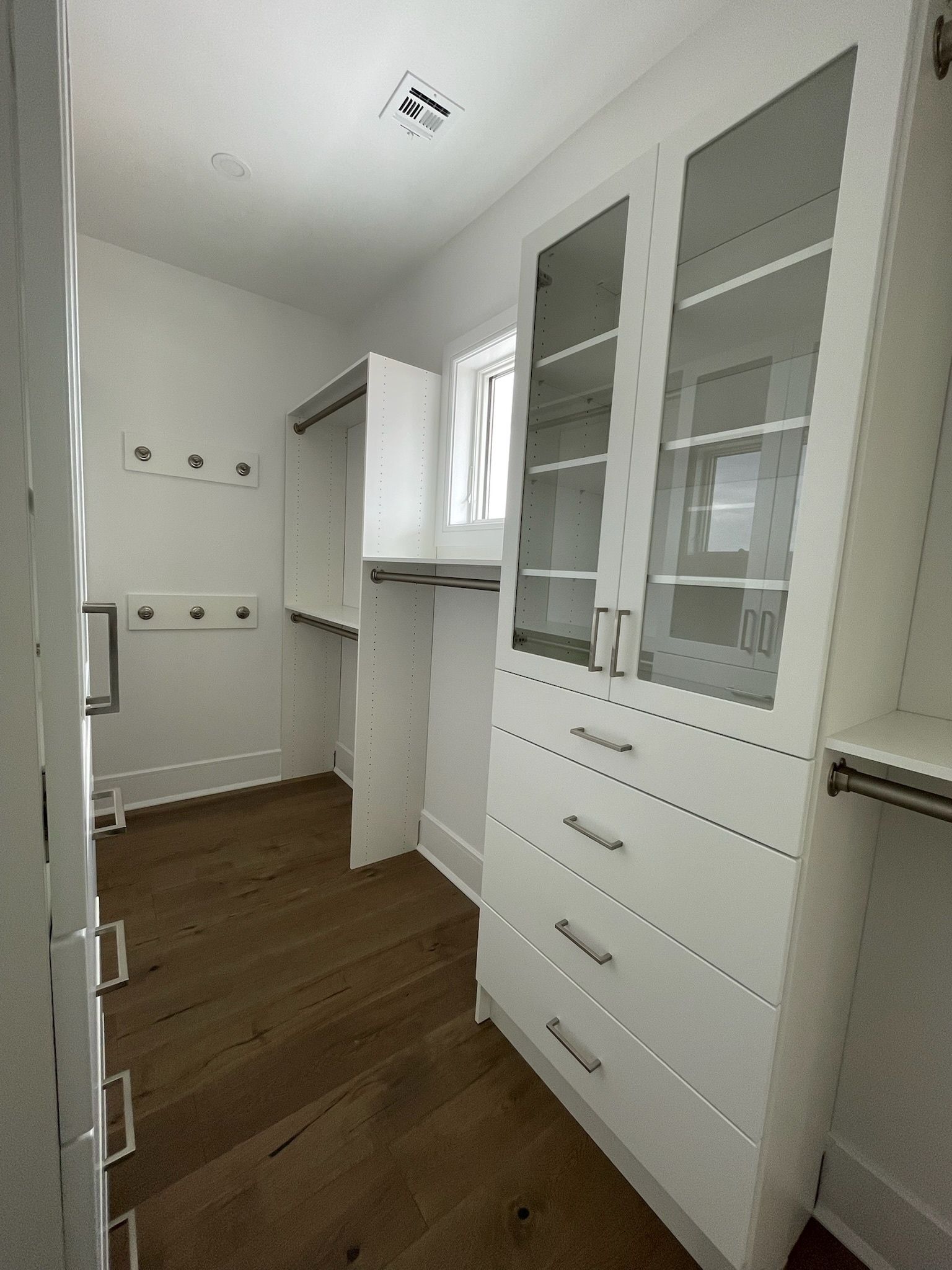 A walk-in closet featuring white built-in storage cabinets, glass-front upper shelves, drawers, and light wood flooring.