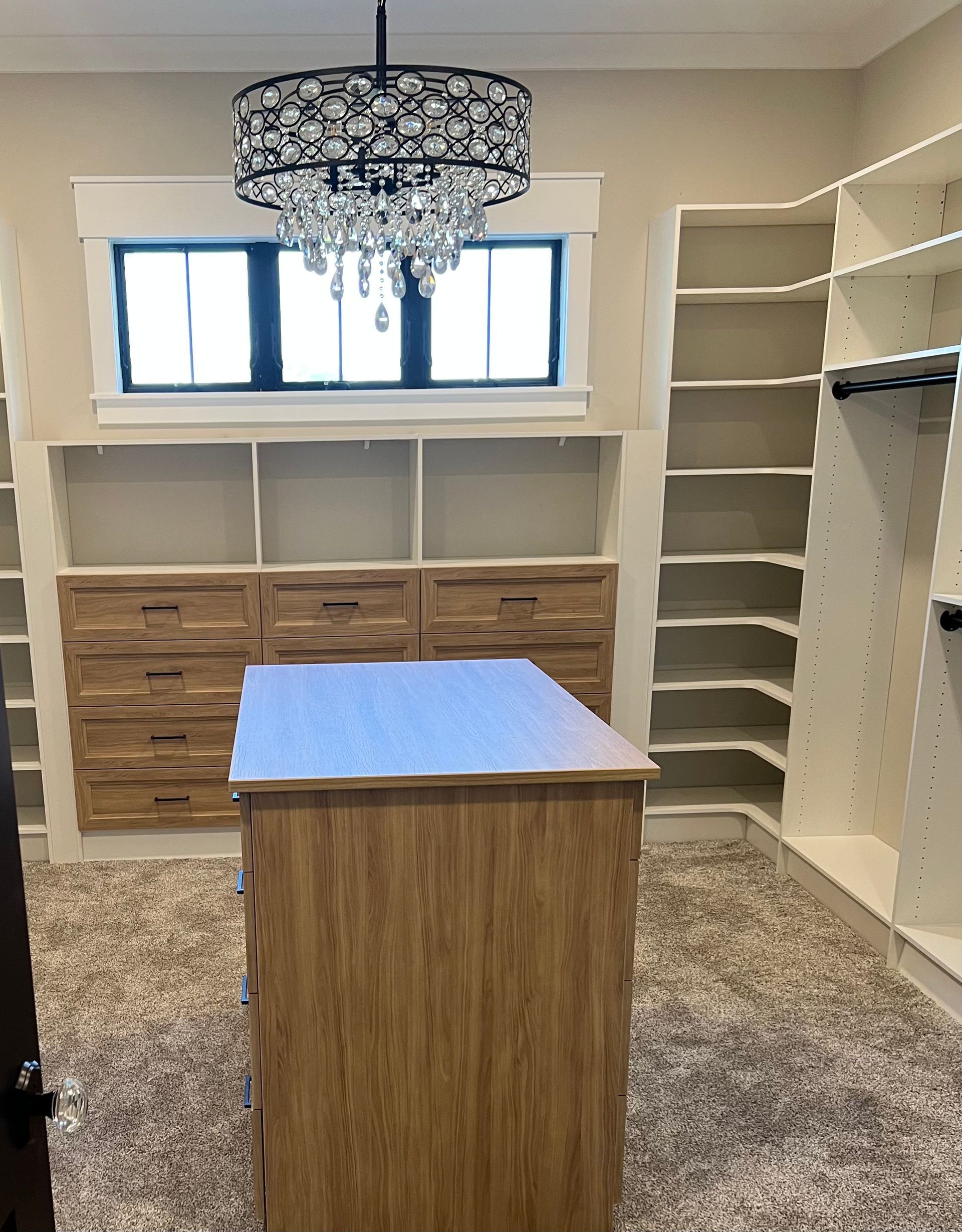 A walk-in closet with tan carpet, light wood built-in drawers and shelves, a central island, and a crystal chandelier.