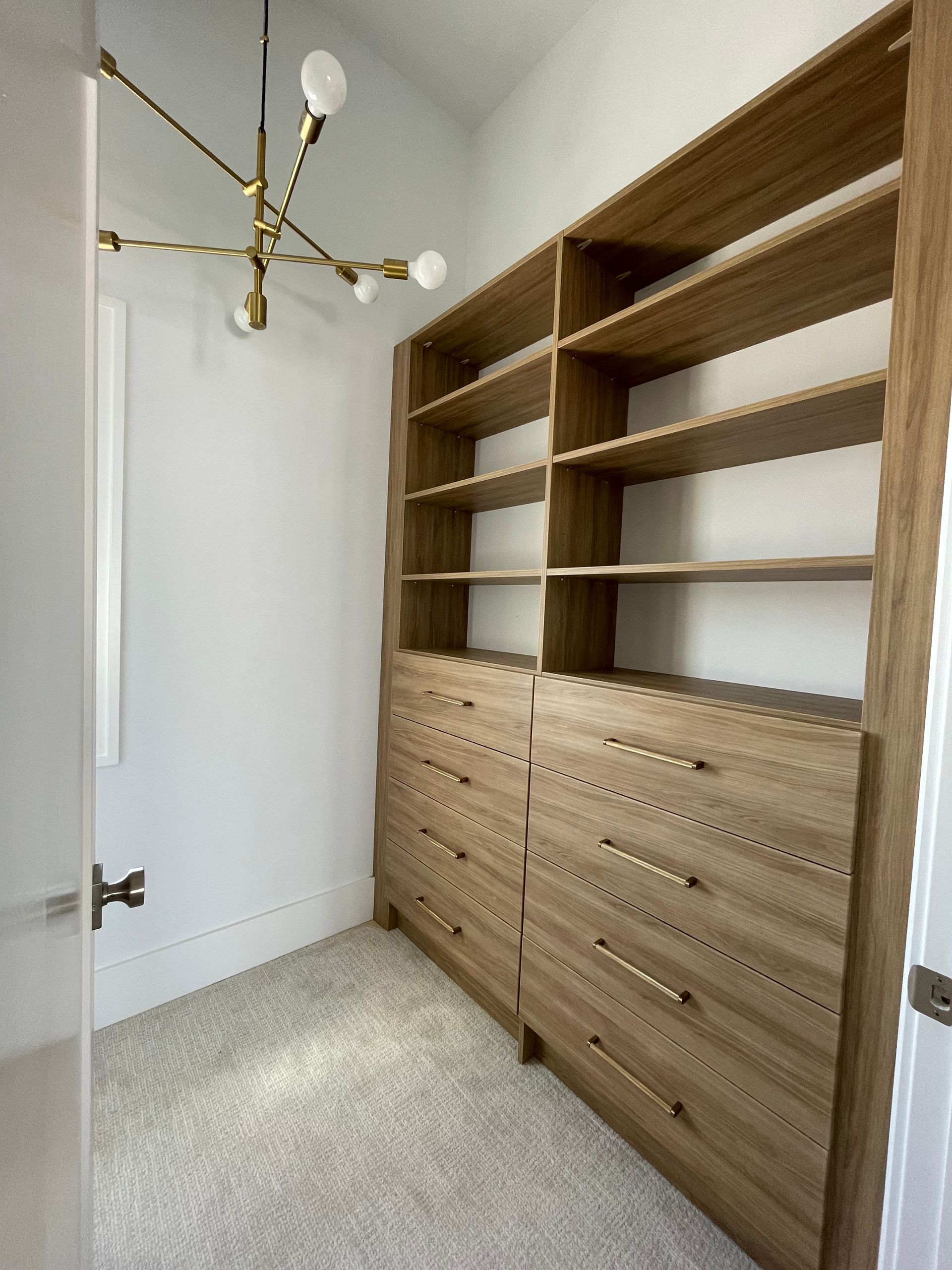A walk-in closet featuring light wood built-in shelves and drawers against white walls, lit by a modern gold chandelier.