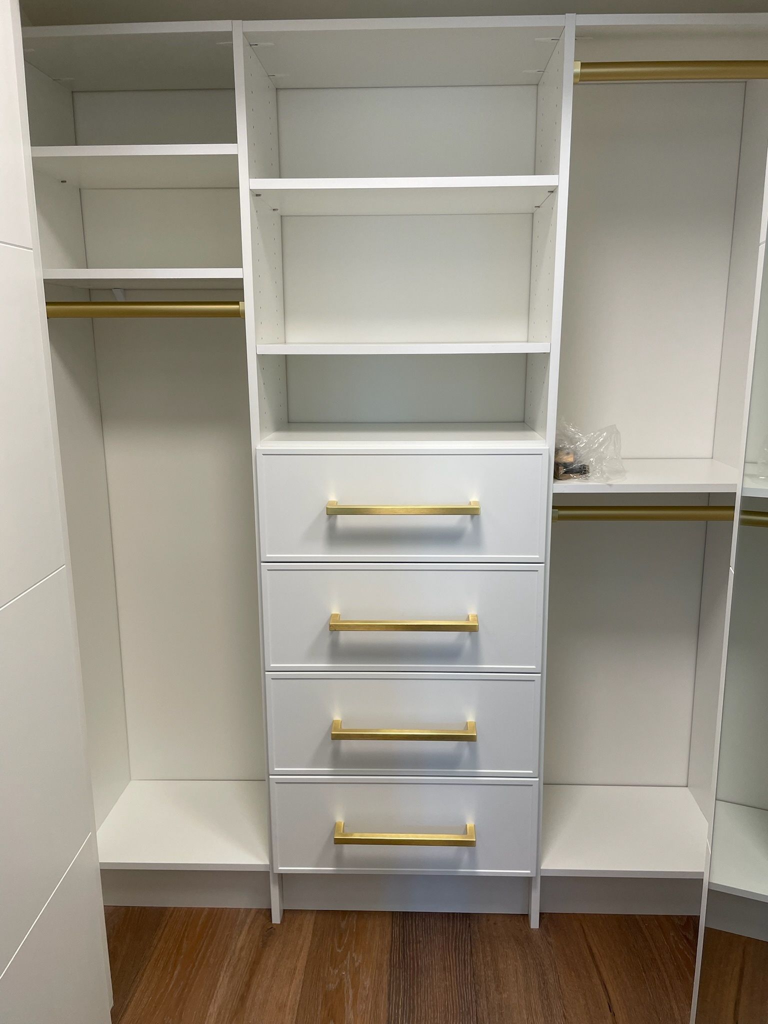 A white closet organizer with three open shelving sections, four drawers with gold handles, and two hanging rods.
