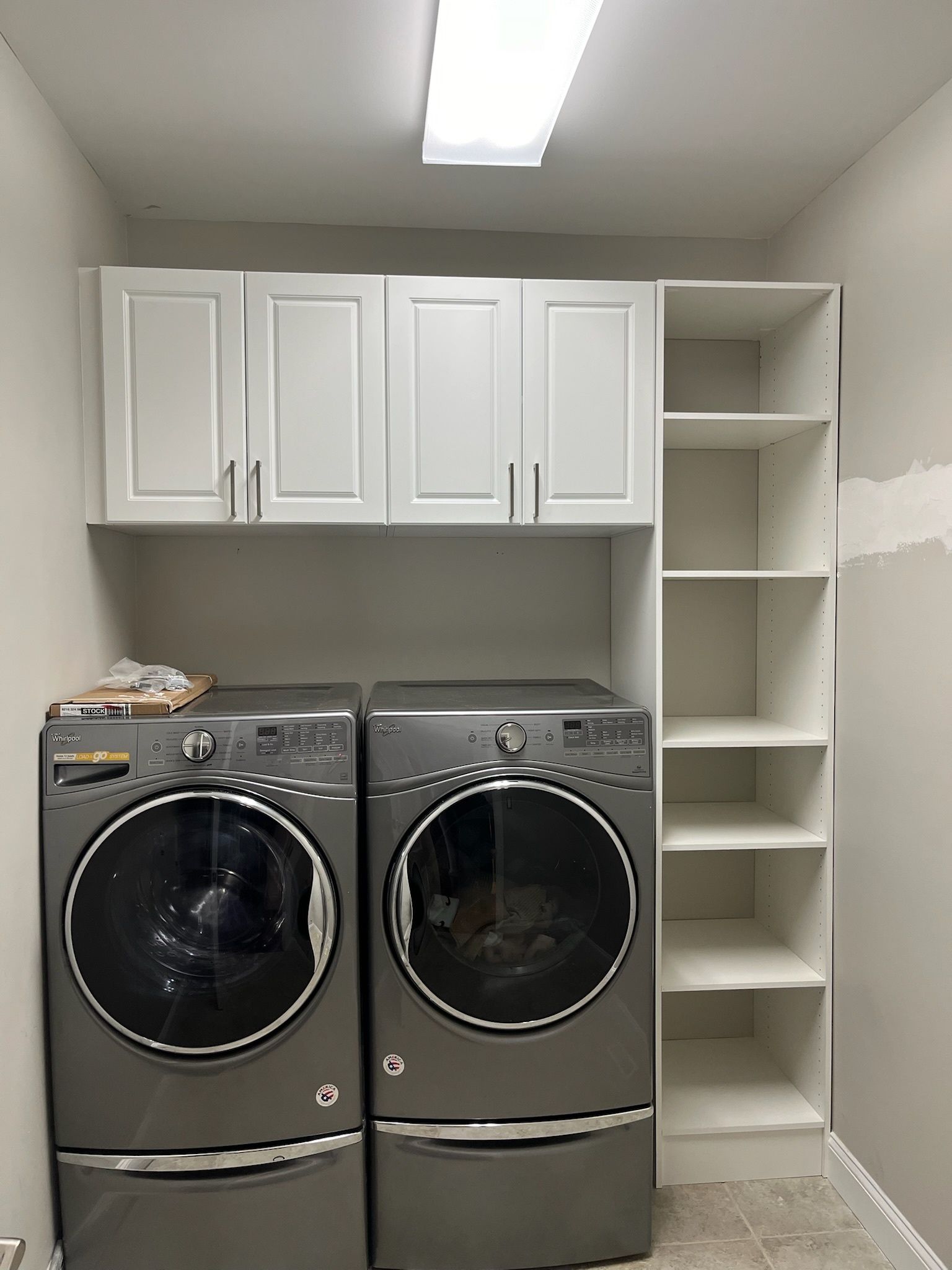 A laundry room featuring two grey front-loading machines under white wall cabinets, next to a tall, white open shelving unit.