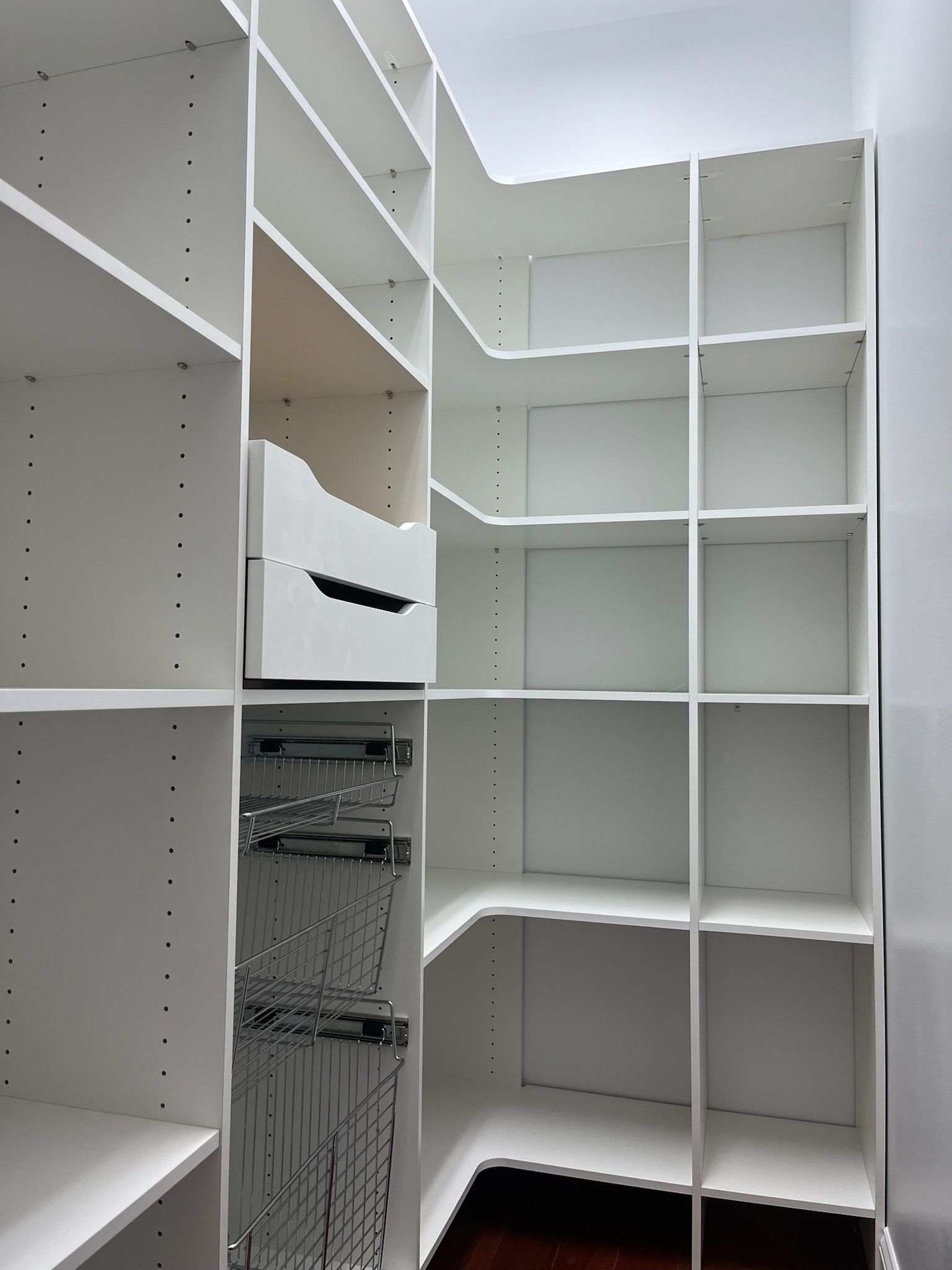 A white, L-shaped walk-in closet shelving system with open storage units, built-in drawers, and metal pull-out baskets.
