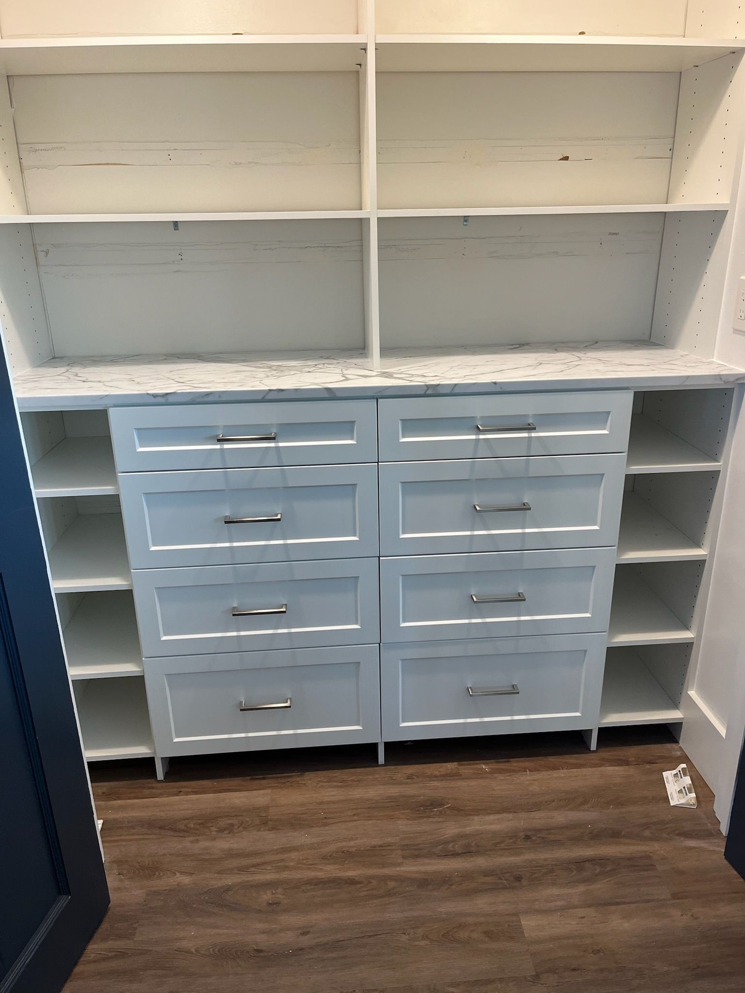 A white built-in closet unit with open shelving on top and sides, featuring eight drawers in the center with silver pulls.