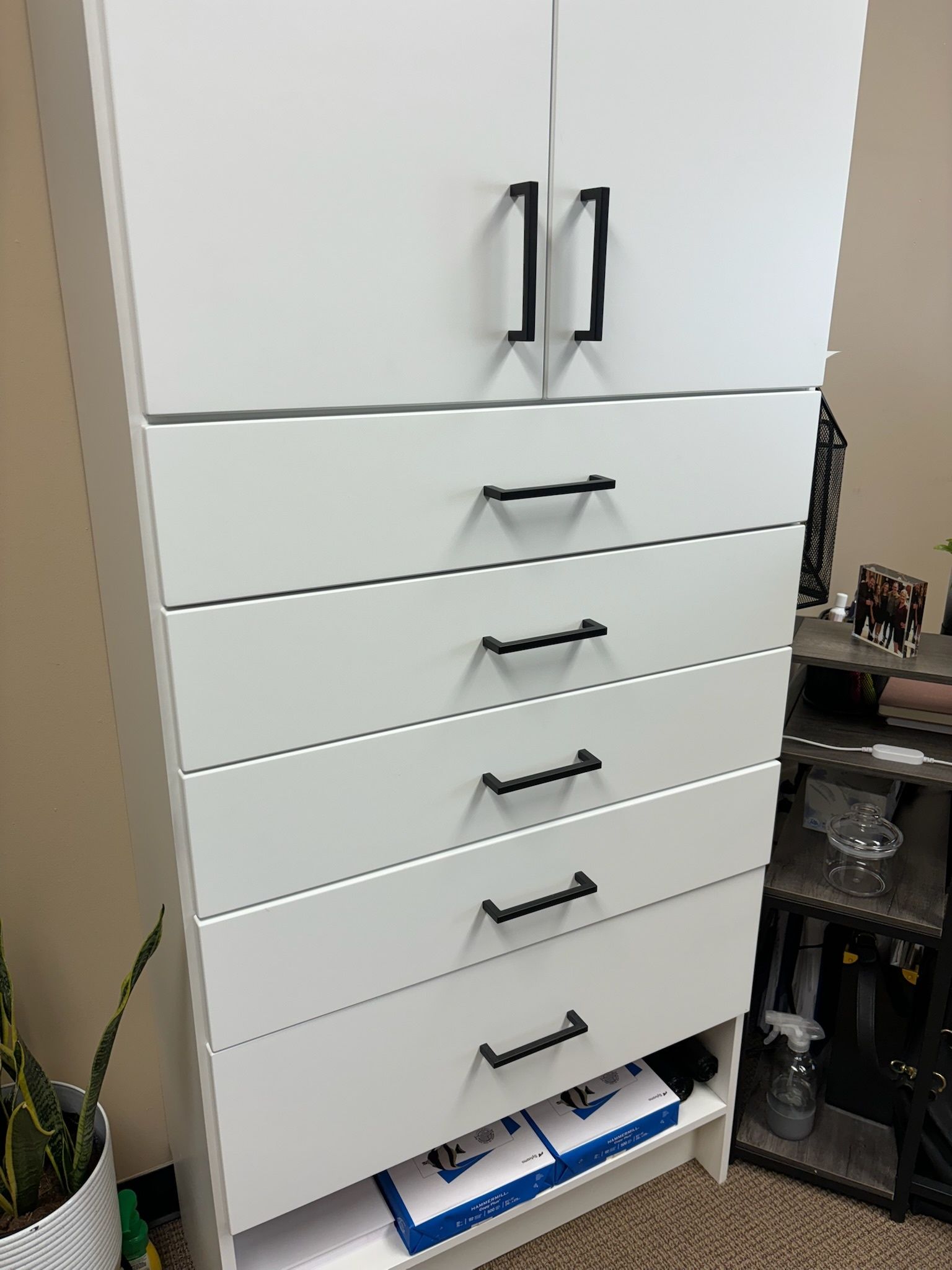 Tall white cabinet with two top doors, five drawers with black handles, and an open bottom shelf in a room.