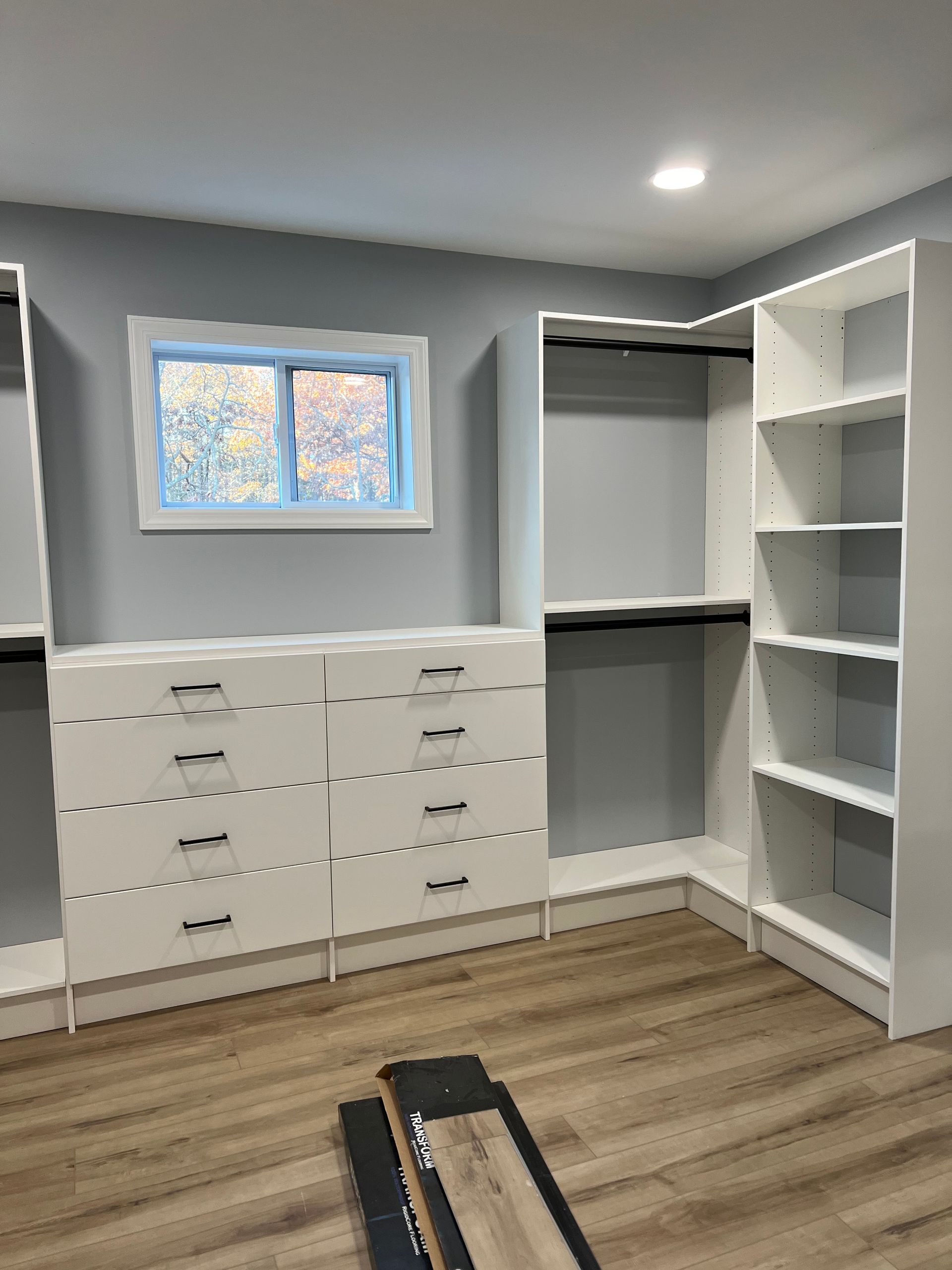 White closet organizers and a dresser installed in a room with grey walls, a small window, and wood-look flooring.