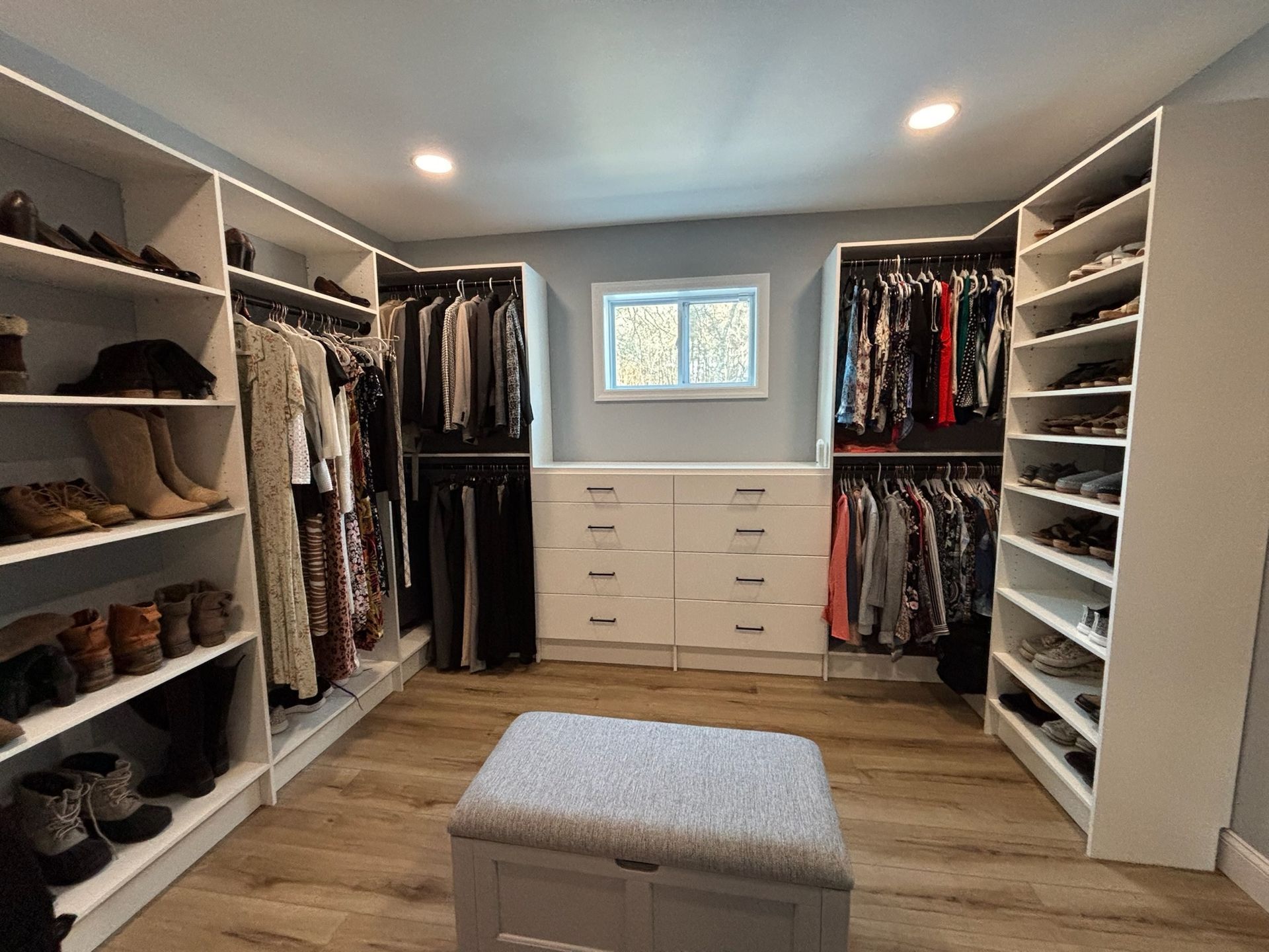 A walk-in closet featuring white shelves, hanging clothes, a center dresser under a window, and a fabric storage ottoman.