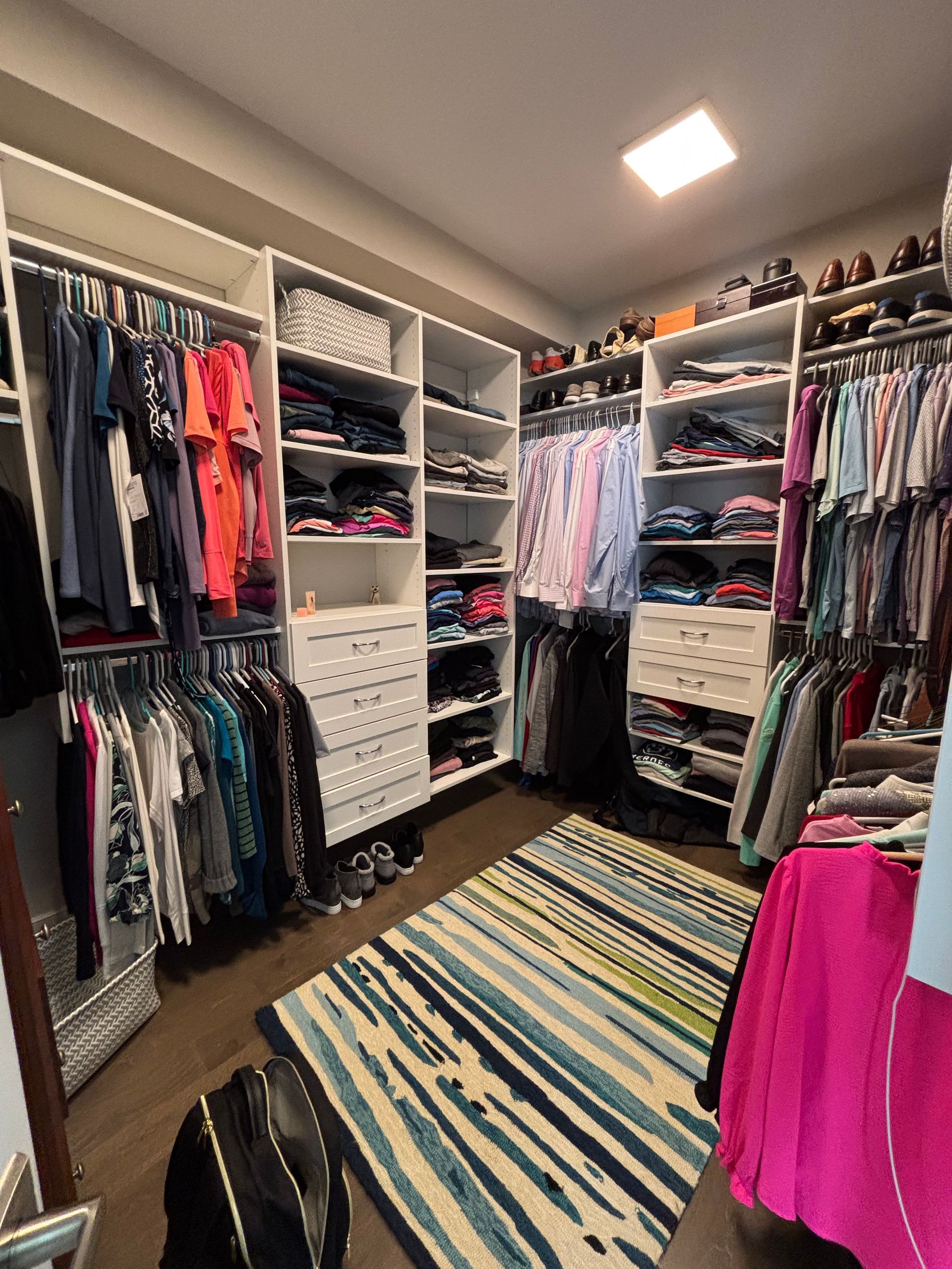 A walk-in closet with white shelving, hanging clothes, folded items, and a striped area rug on the floor.