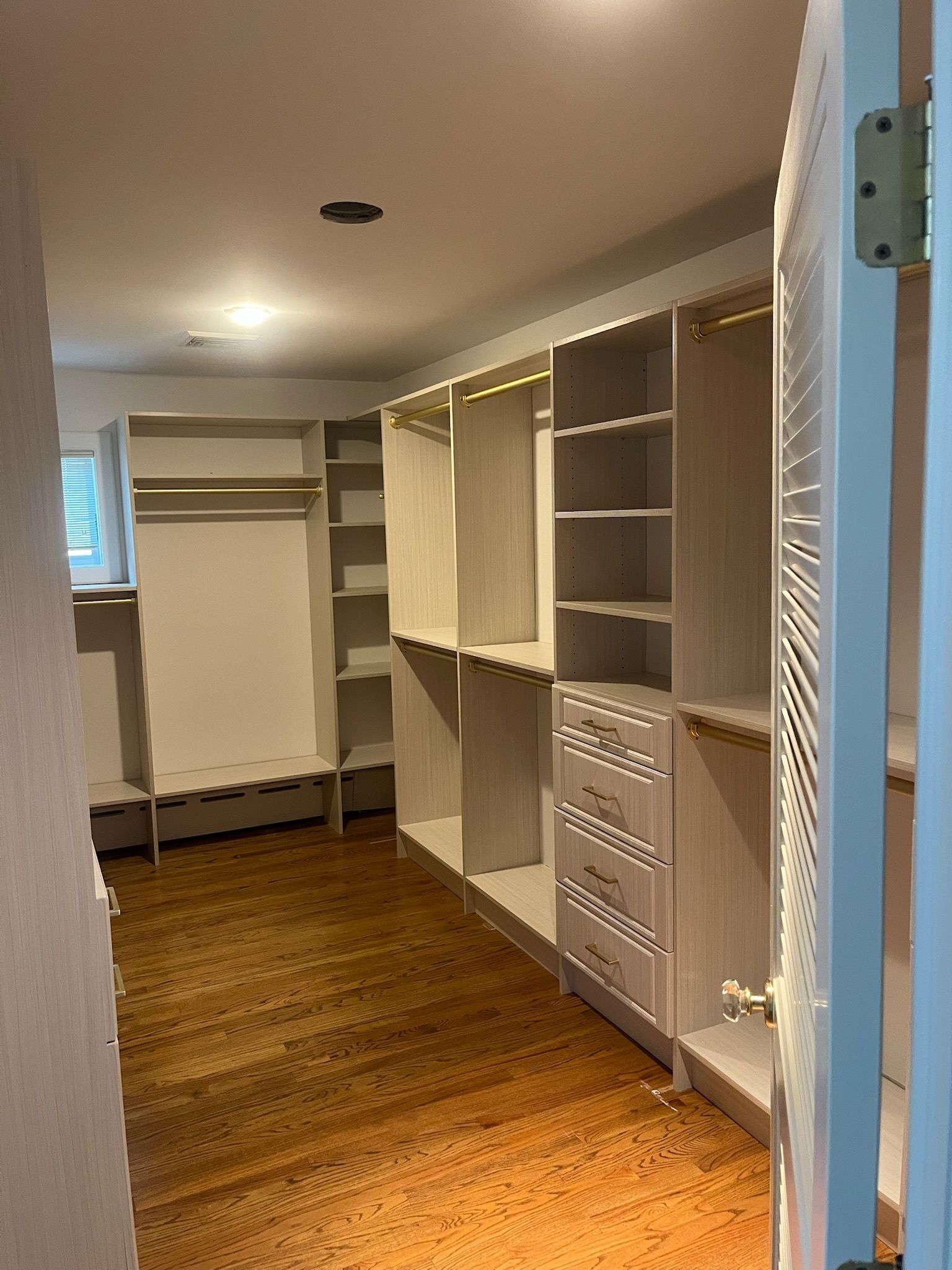A walk-in closet featuring light-colored wooden shelving units, hanging rods, and drawers, viewed from a doorway.