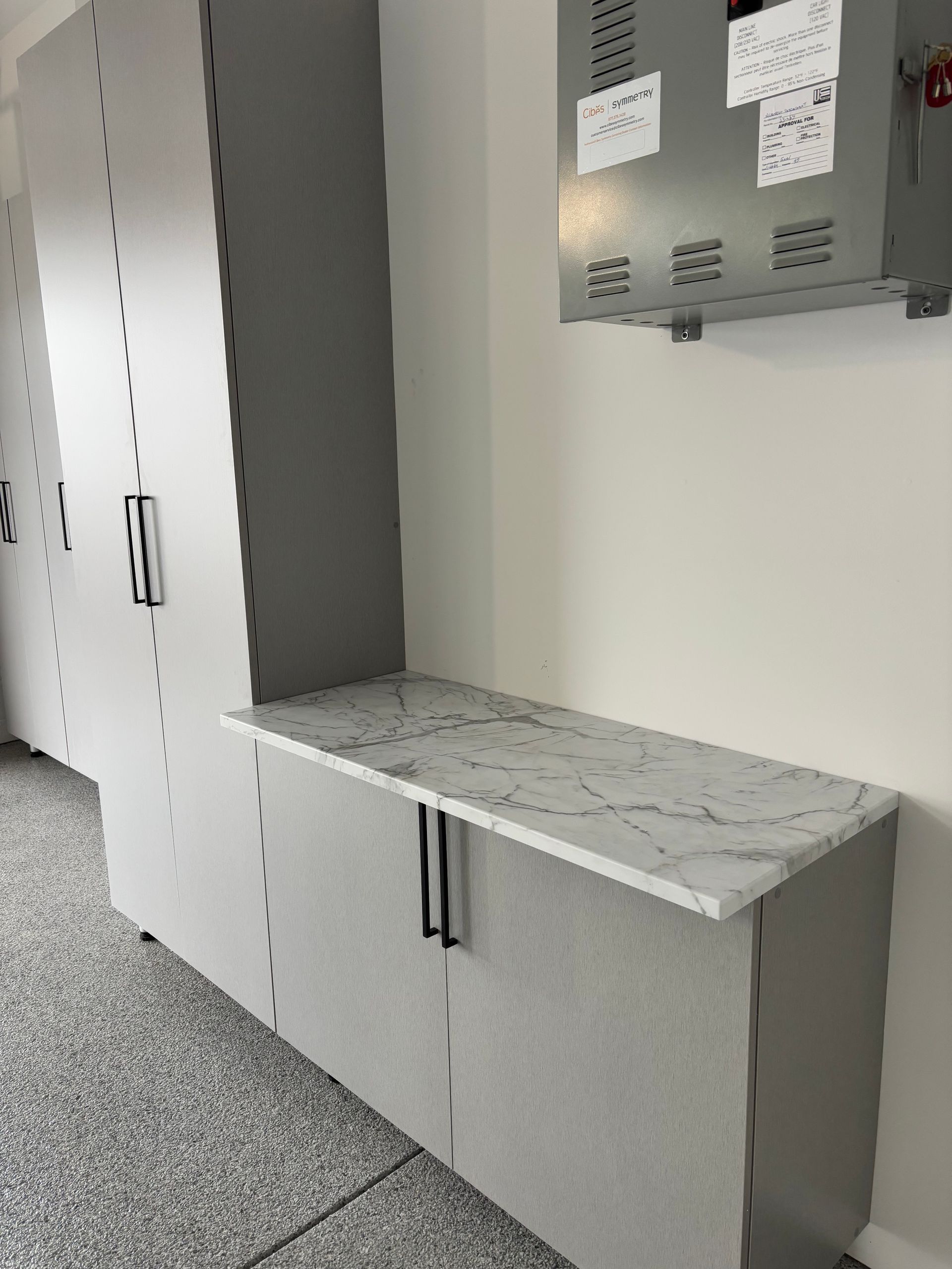A tall gray storage locker next to a base cabinet with a white marble-patterned countertop in a garage setting.