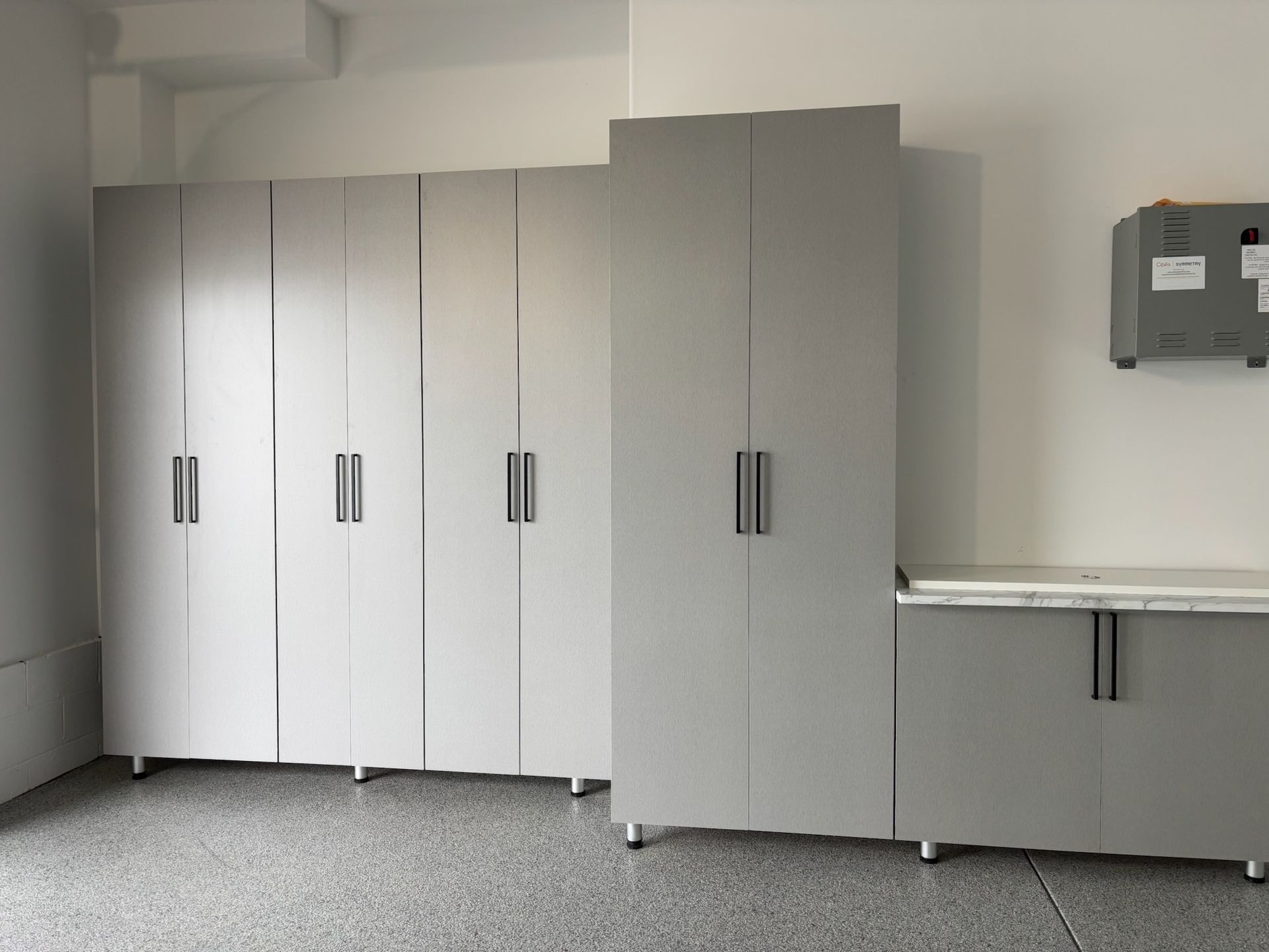 A row of modern light-gray garage storage cabinets with silver handles installed against a white wall on a speckled floor.