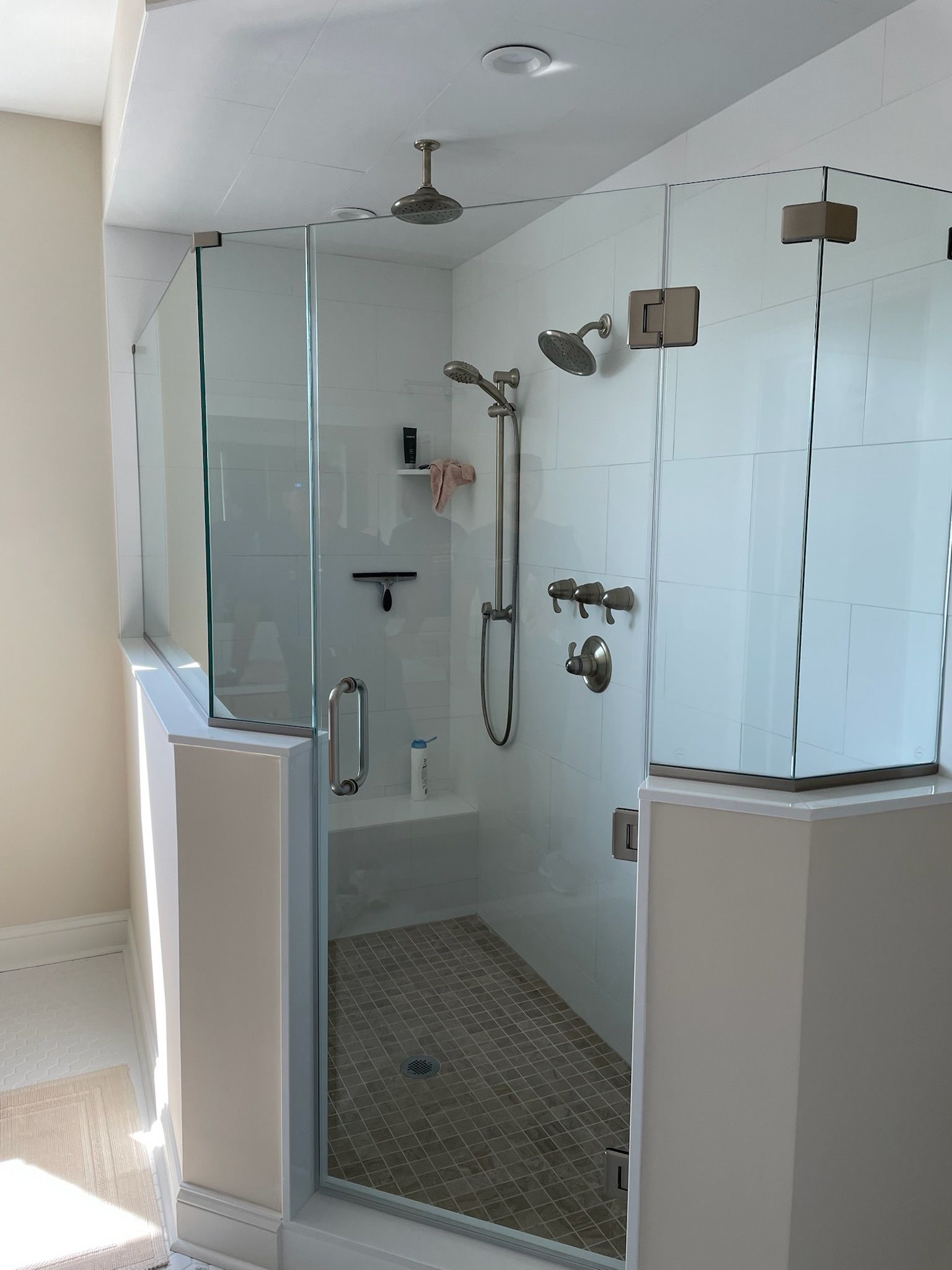 A modern walk-in shower with a glass door, white tiled walls, a stone-patterned floor, and multiple shower heads.