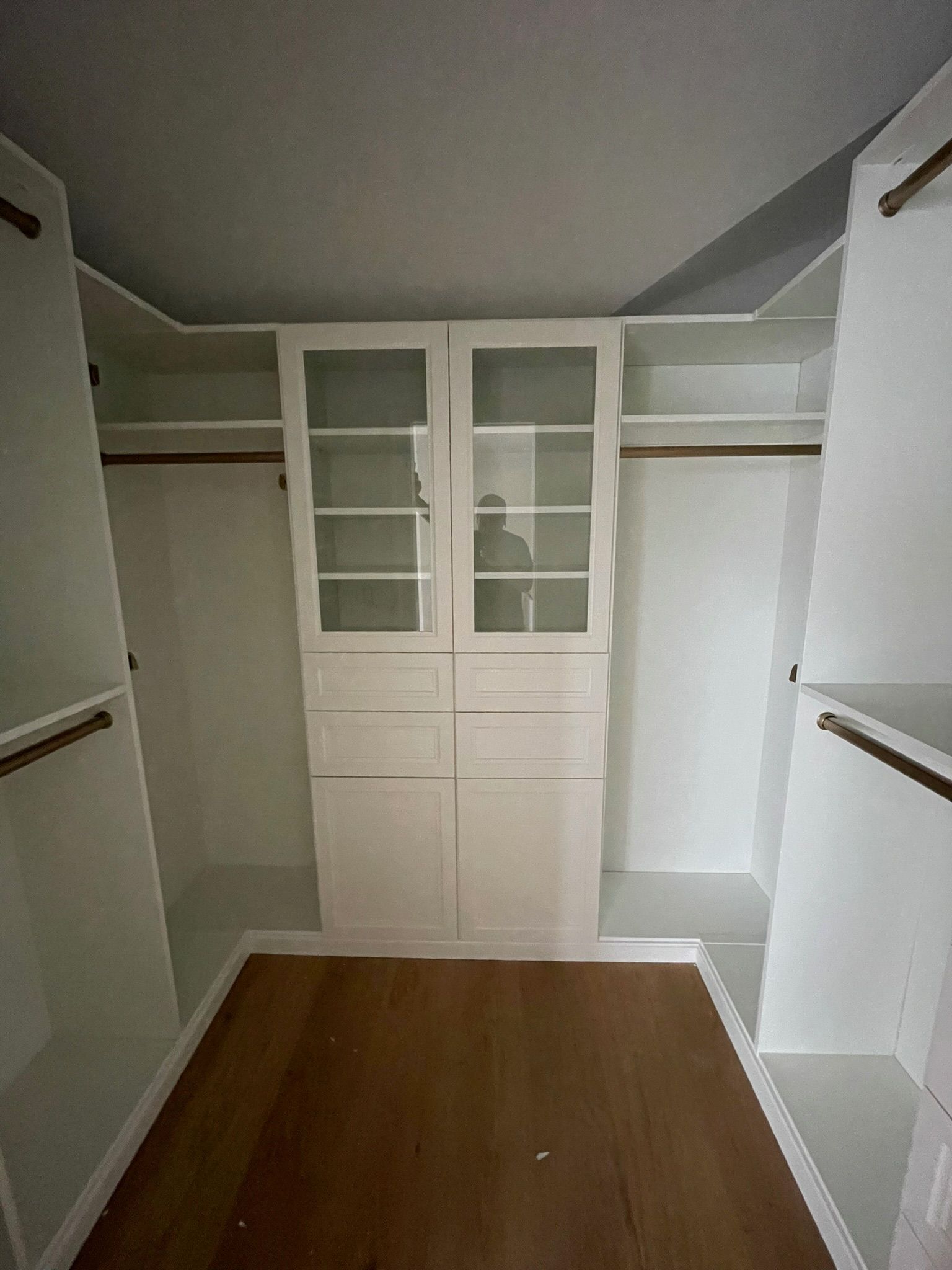 A walk-in closet featuring white wooden cabinetry, glass-front upper shelves, drawers, and hanging rods on light wood floors.