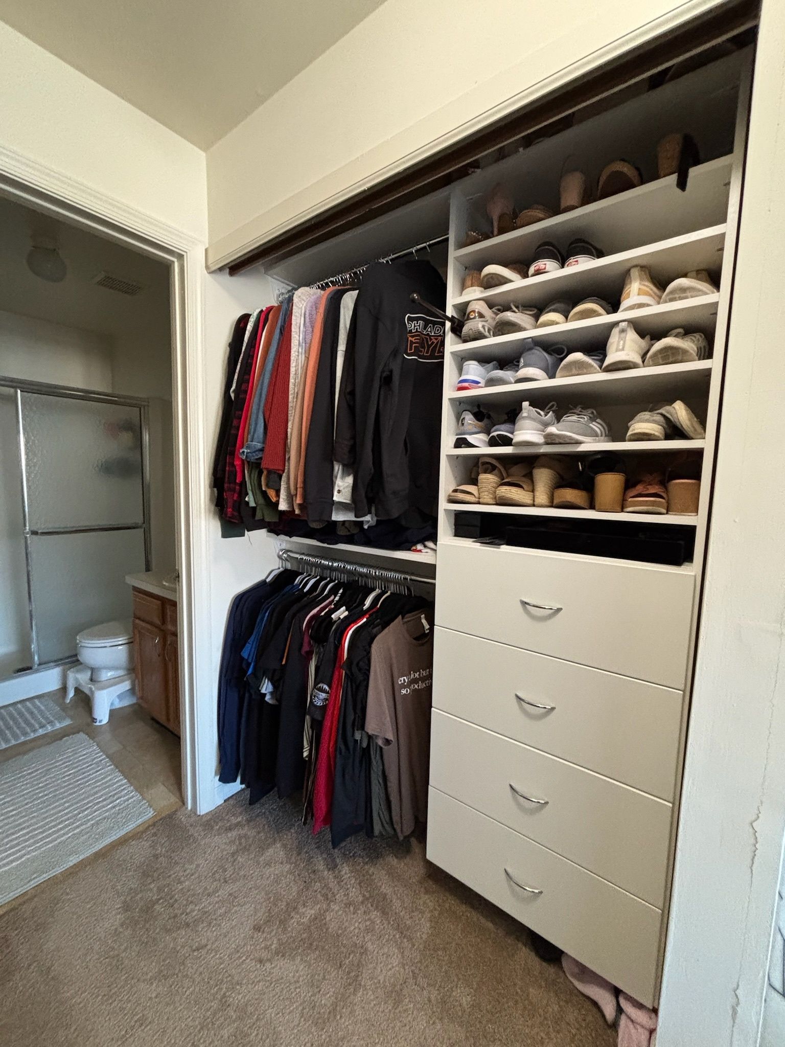 A closet with hanging clothes on rods, a vertical shoe rack, and four drawers, adjacent to a bathroom.