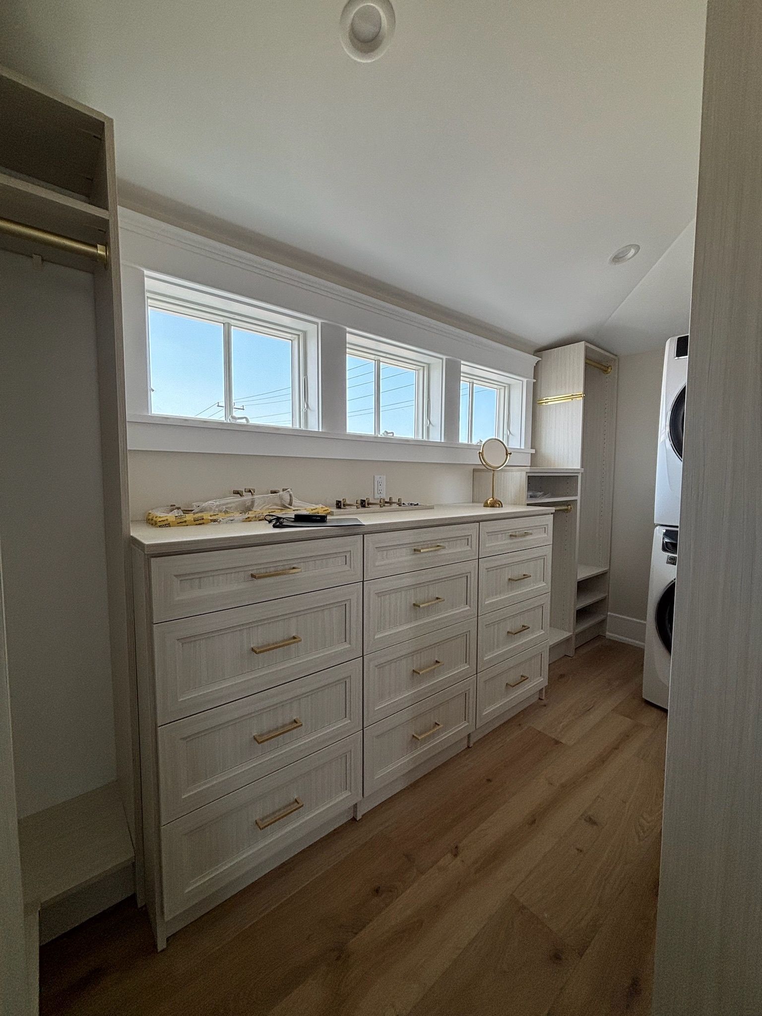 A bright, modern closet with light wood flooring, white drawers under windows, open shelving, and a laundry appliance.