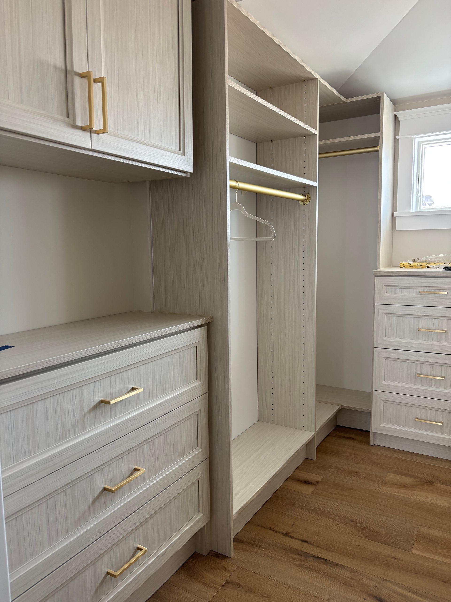 Light-wood closet system with gold hardware, including drawers, shelves, and a hanging rod in a room with wood floors.