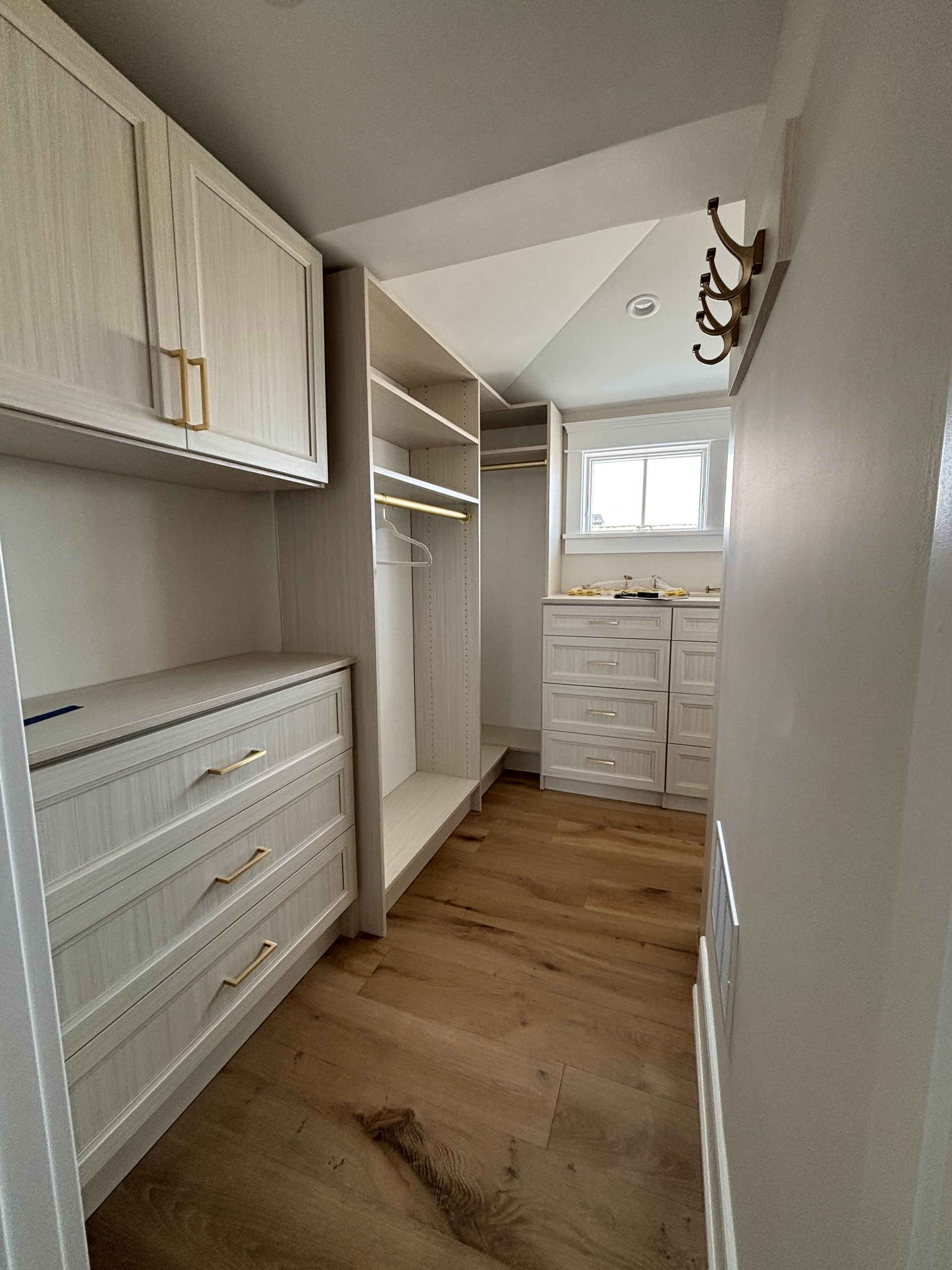 A walk-in closet with light wood-grained cabinets, drawers, open shelving, a wooden floor, and a window at the end.