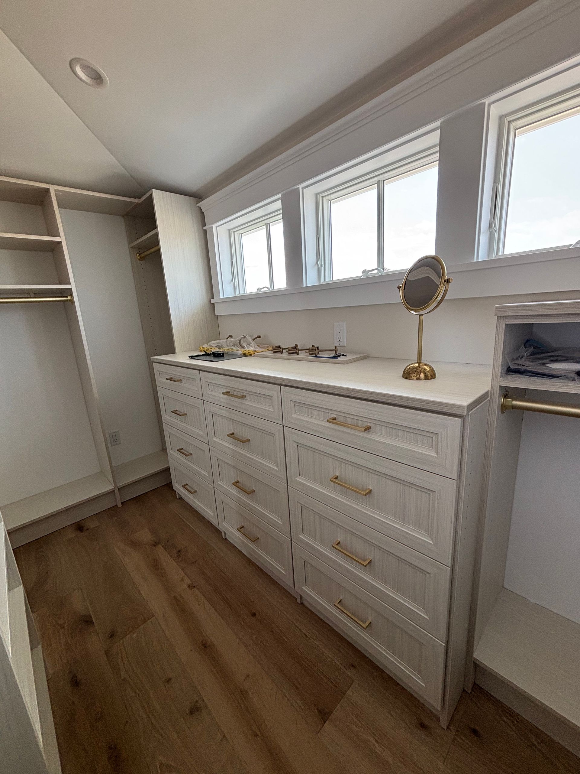 A walk-in closet with cream-colored cabinetry, drawers, and shelving, featuring a makeup mirror and wood flooring.