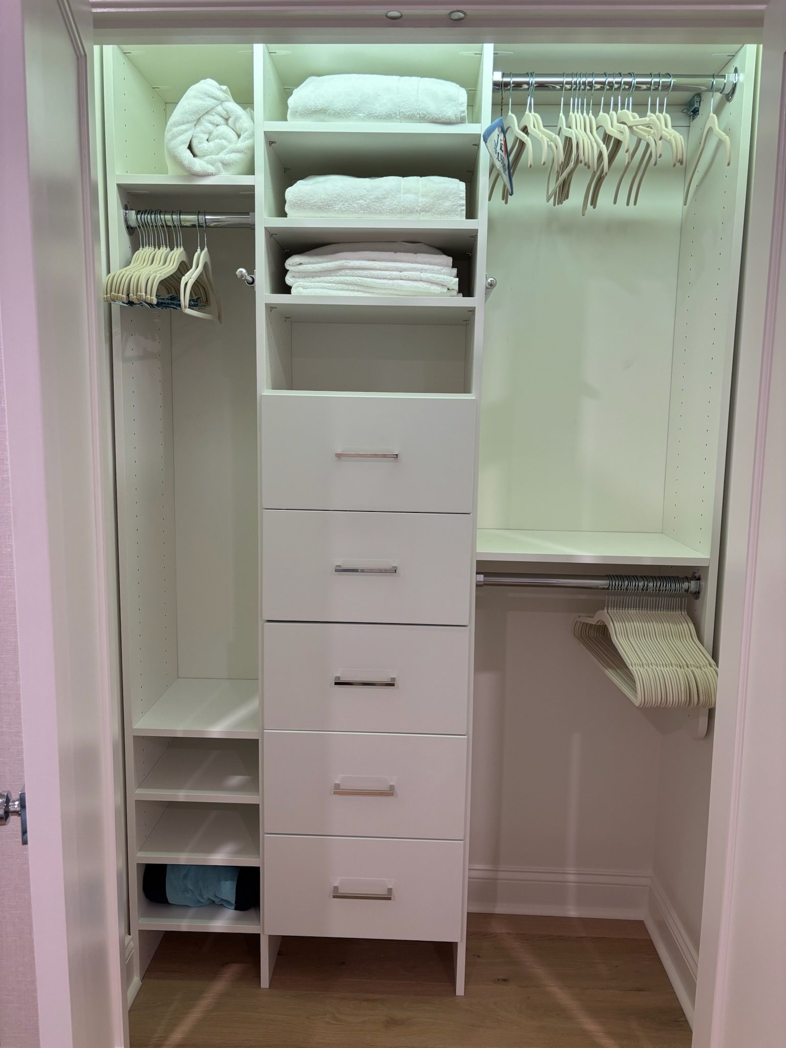 A white closet interior featuring hanging rods, open shelving, and a column of five drawers, with towels on the shelves.