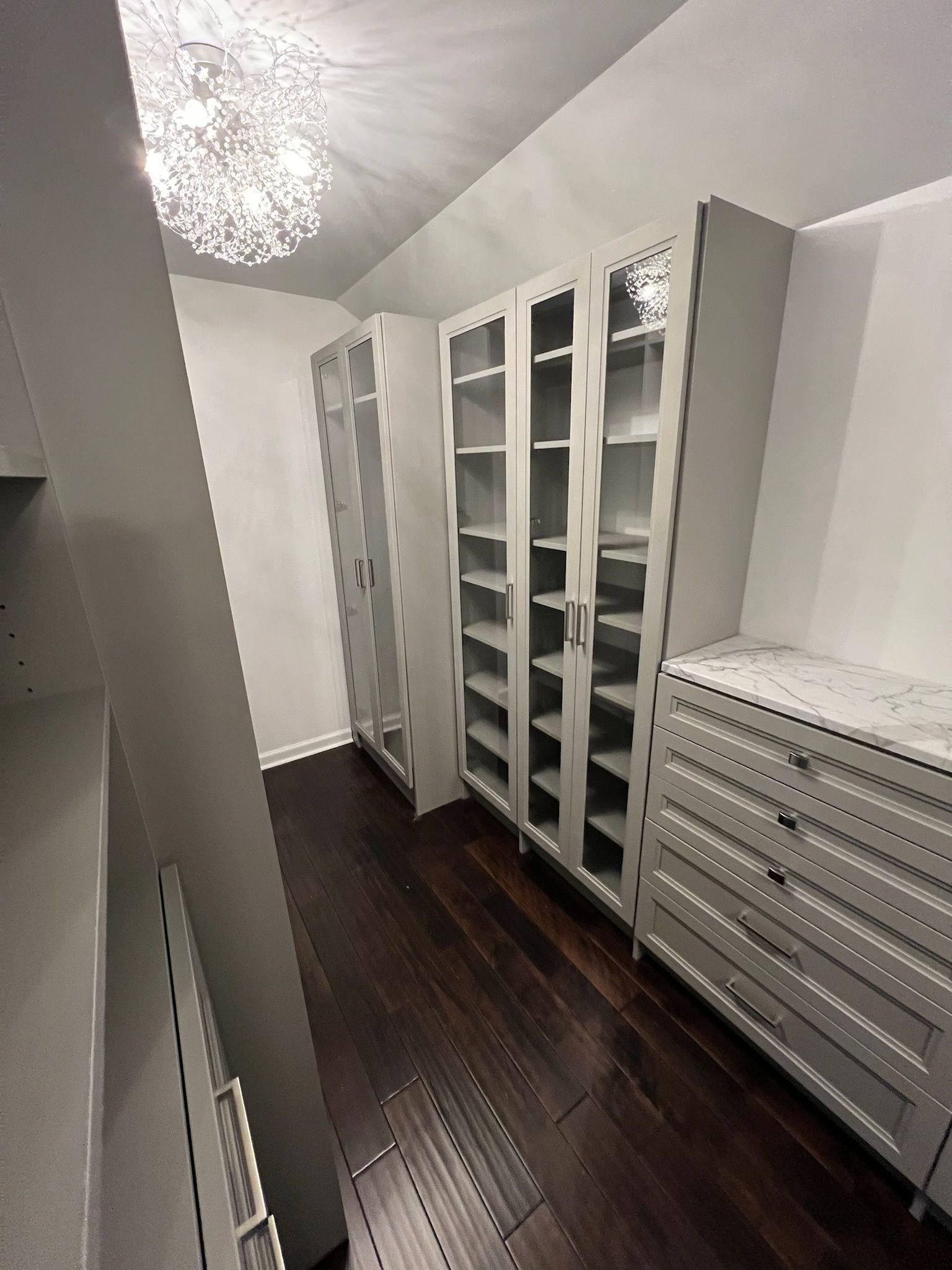 A narrow walk-in closet featuring light grey floor-to-ceiling glass cabinets, a matching dresser, and dark hardwood floors.